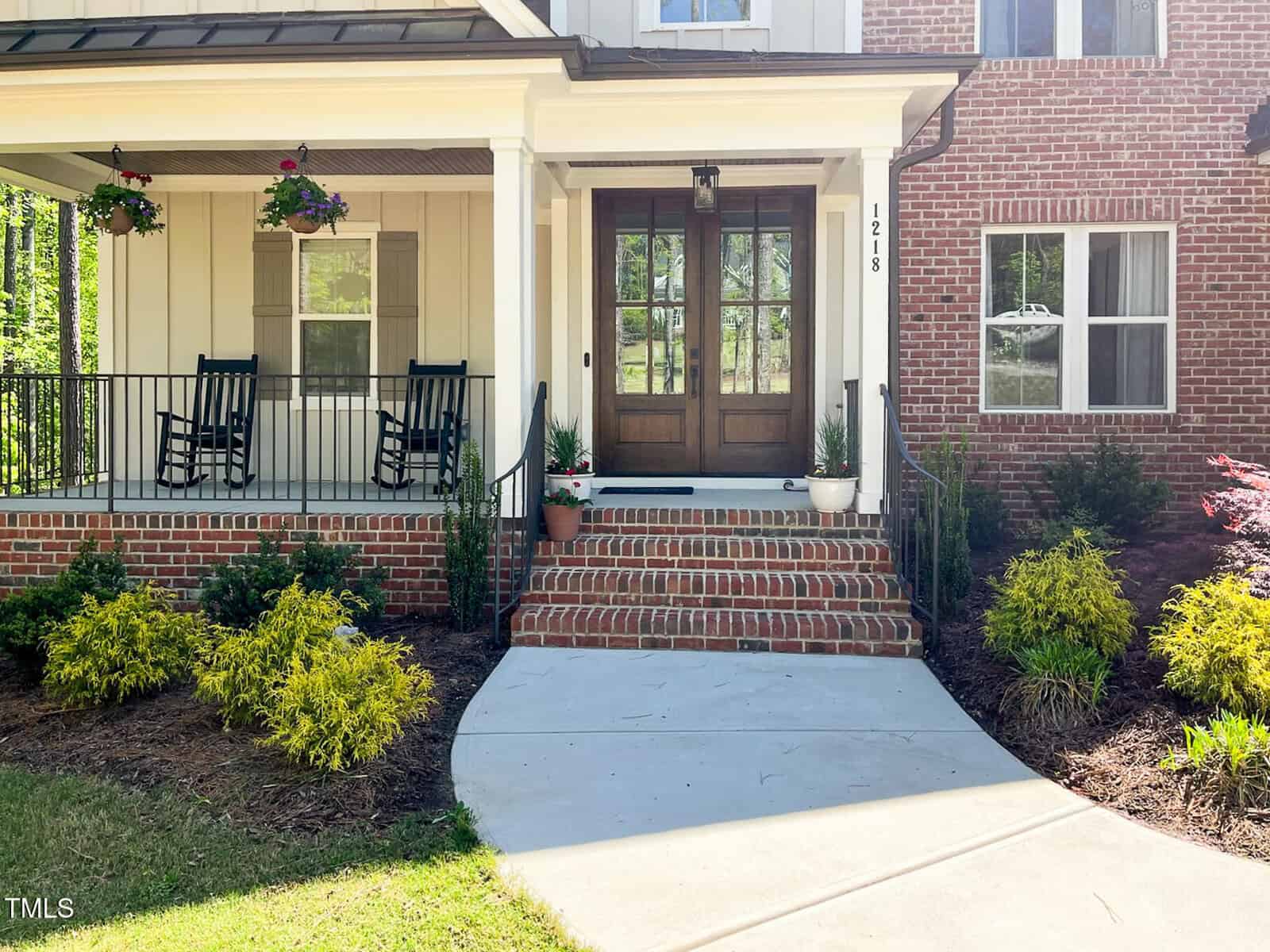 A house entrance with brick steps, a wooden double front door, two black rocking chairs on a covered porch, hanging flower baskets, potted plants, and landscaping with shrubs and bushes.