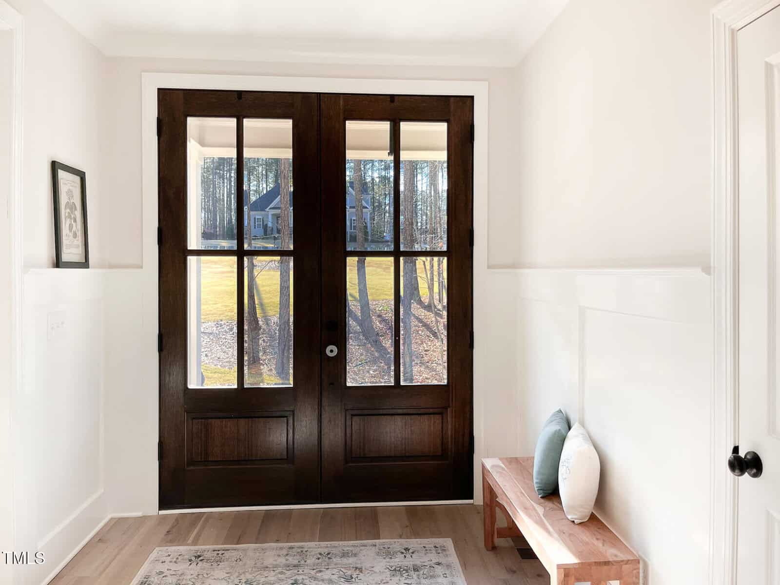 A bright entryway with large double wooden doors featuring glass panels. A wooden bench with two throw pillows sits to the right, and a framed picture hangs on the left wall. Sunlight streams through the doors.