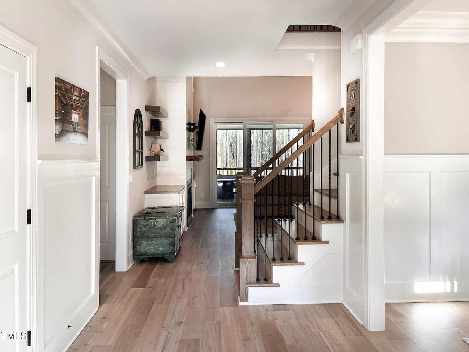 A hallway with light wood floors leads to a living area with large windows. A staircase with wooden steps and black railing is on the right. White walls and trim, minimal decor, and a green chest are visible.