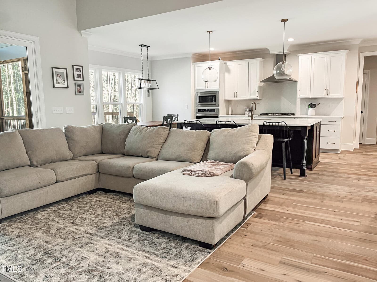 A spacious open-plan living room and kitchen area featuring a beige sectional sofa, a patterned rug, bar stools at a kitchen island, and white cabinets with stainless steel appliances. Natural light fills the room.