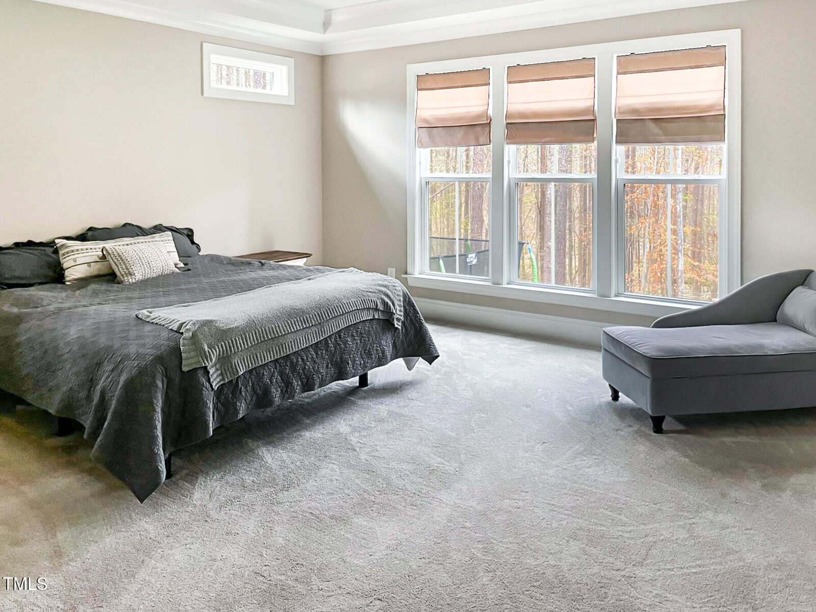 A spacious bedroom with light-colored carpet, a dark bed with pillows and a throw blanket, a gray chaise lounge by large windows with beige shades, and neutral walls with a small upper window.