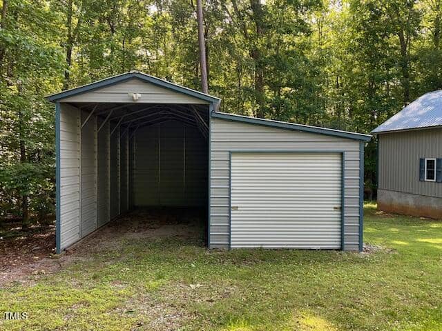 A metal carport with an attached garage featuring a roll-up door is situated on a grassy area near a wooded lot. Another building with a metal roof is partially visible on the right.