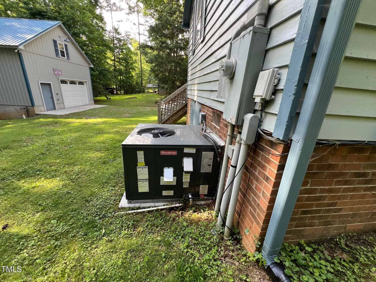 An outdoor HVAC unit is installed next to a brick house wall with utility meters and pipes attached. A detached garage and trees are visible in the background on a grassy lawn.
