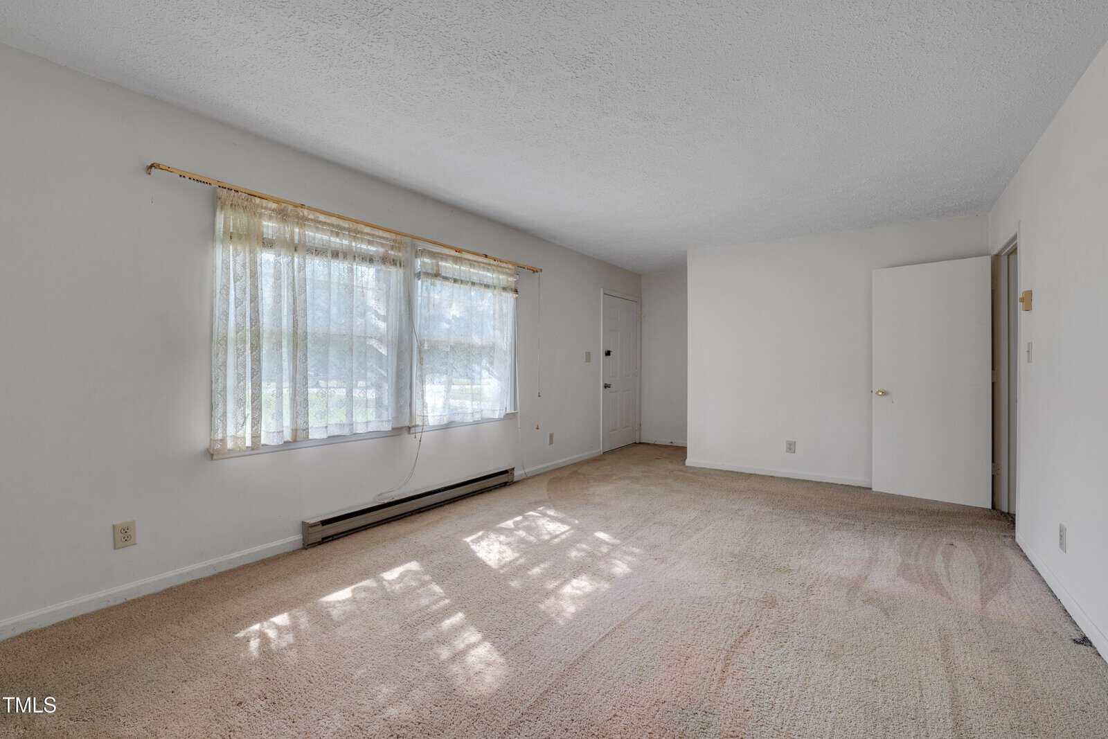 An empty room with beige carpet, white walls, a large window with sheer curtains, a baseboard heater below the window, and an open white door leading to another area. Natural light fills the room.