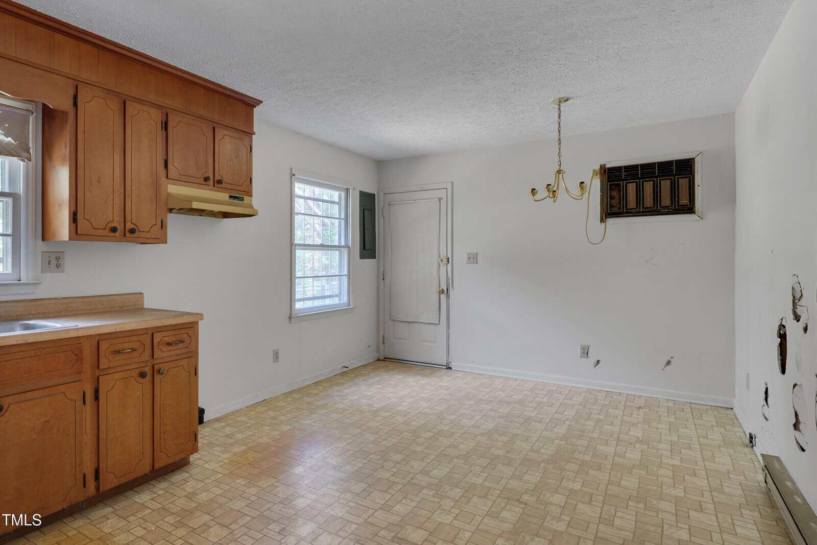 An empty kitchen with wooden cabinets, a beige countertop, linoleum flooring, a window, a wall-mounted air conditioning unit, a ceiling light fixture missing shades, and exposed plumbing on one wall.