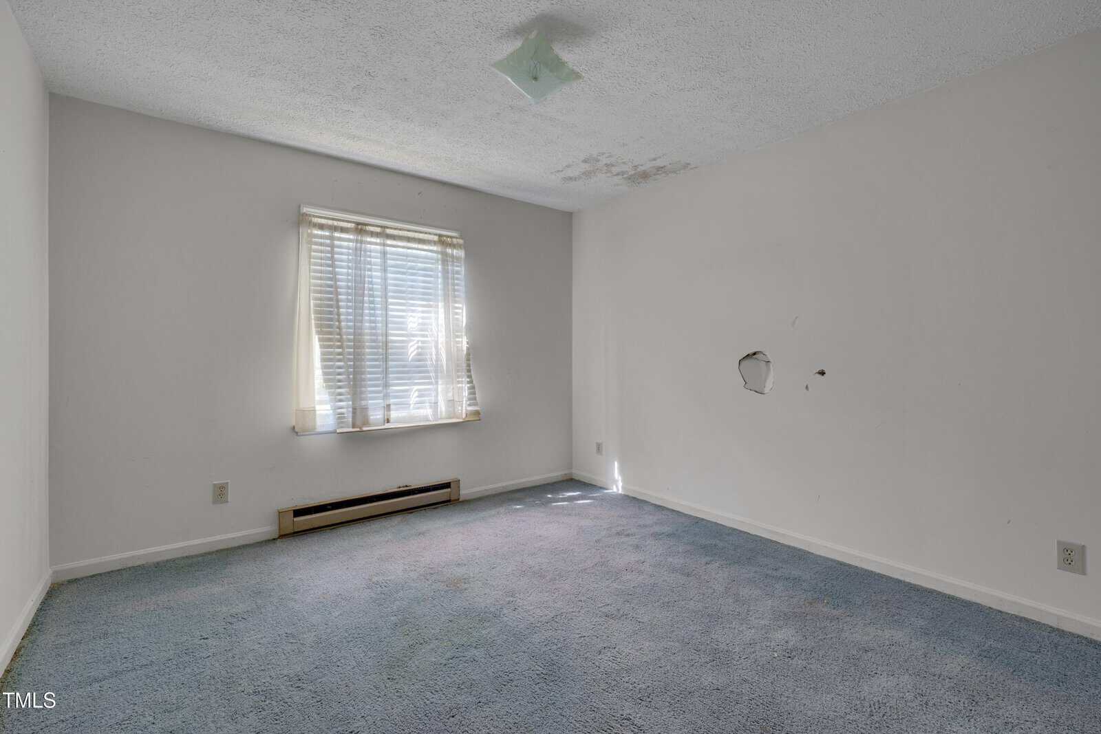 Empty room with white walls, a window with blinds letting in sunlight, blue carpeted floor, a heater under the window, a ceiling light fixture, water stains on the ceiling, and a hole in the right wall.