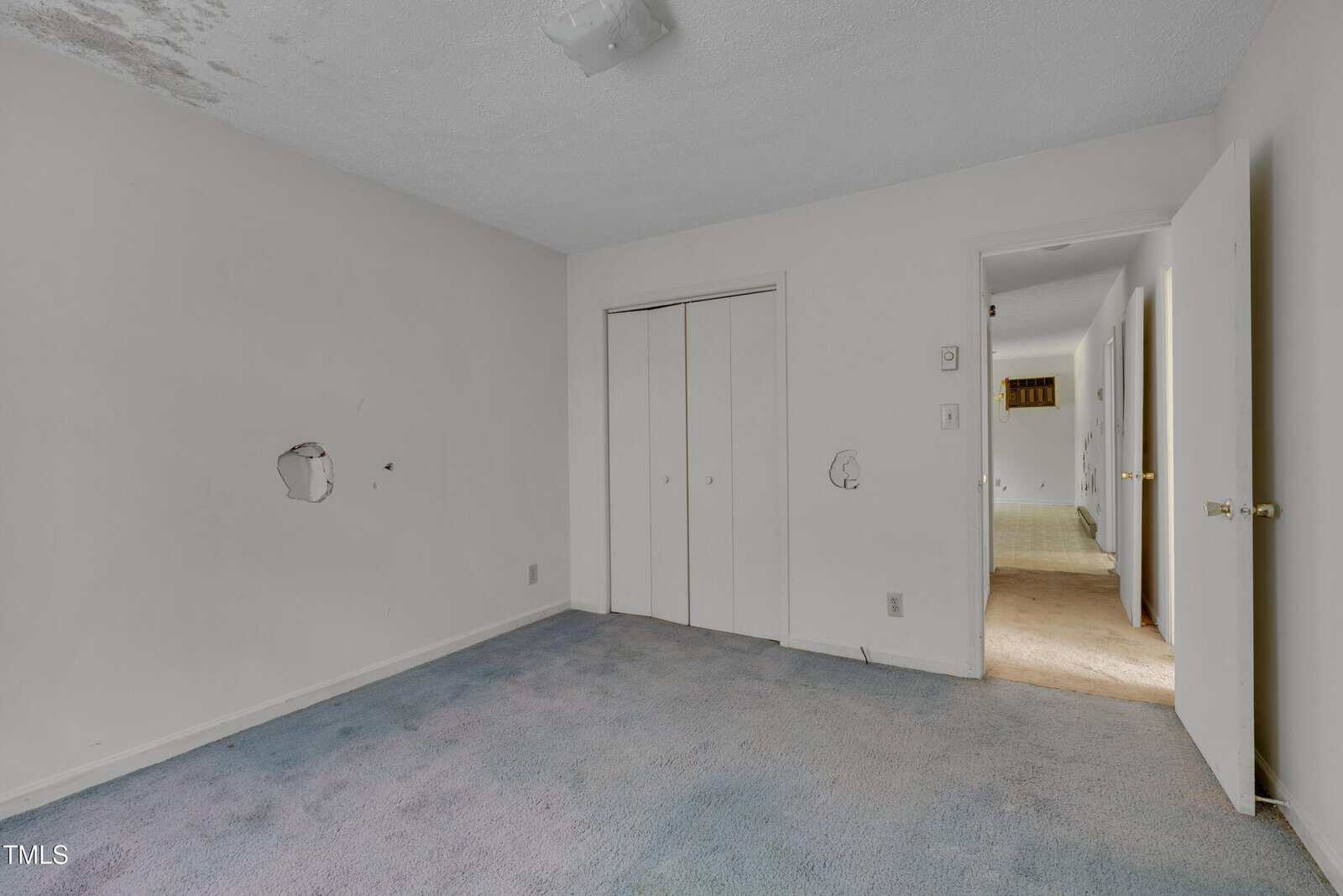A small empty room with white walls, two holes in the wall, a white closet with double doors, a light fixture on the ceiling, and worn blue carpet. An open door leads to a hallway with beige carpeting.