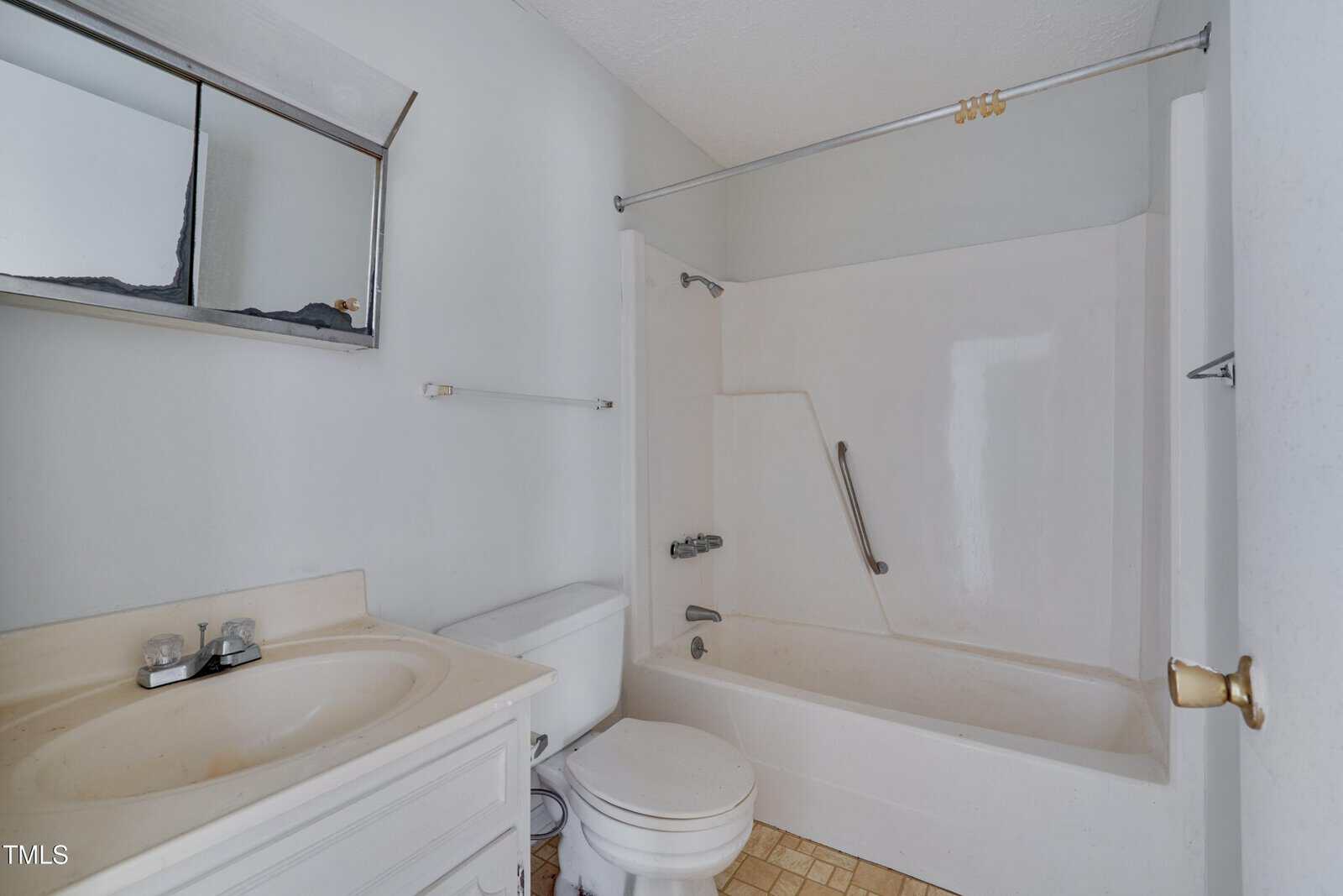 A small bathroom with a white vanity and sink, a toilet, and a bathtub-shower combination with a grab bar. The floor has beige tiles and there is a wall-mounted mirror above the sink.