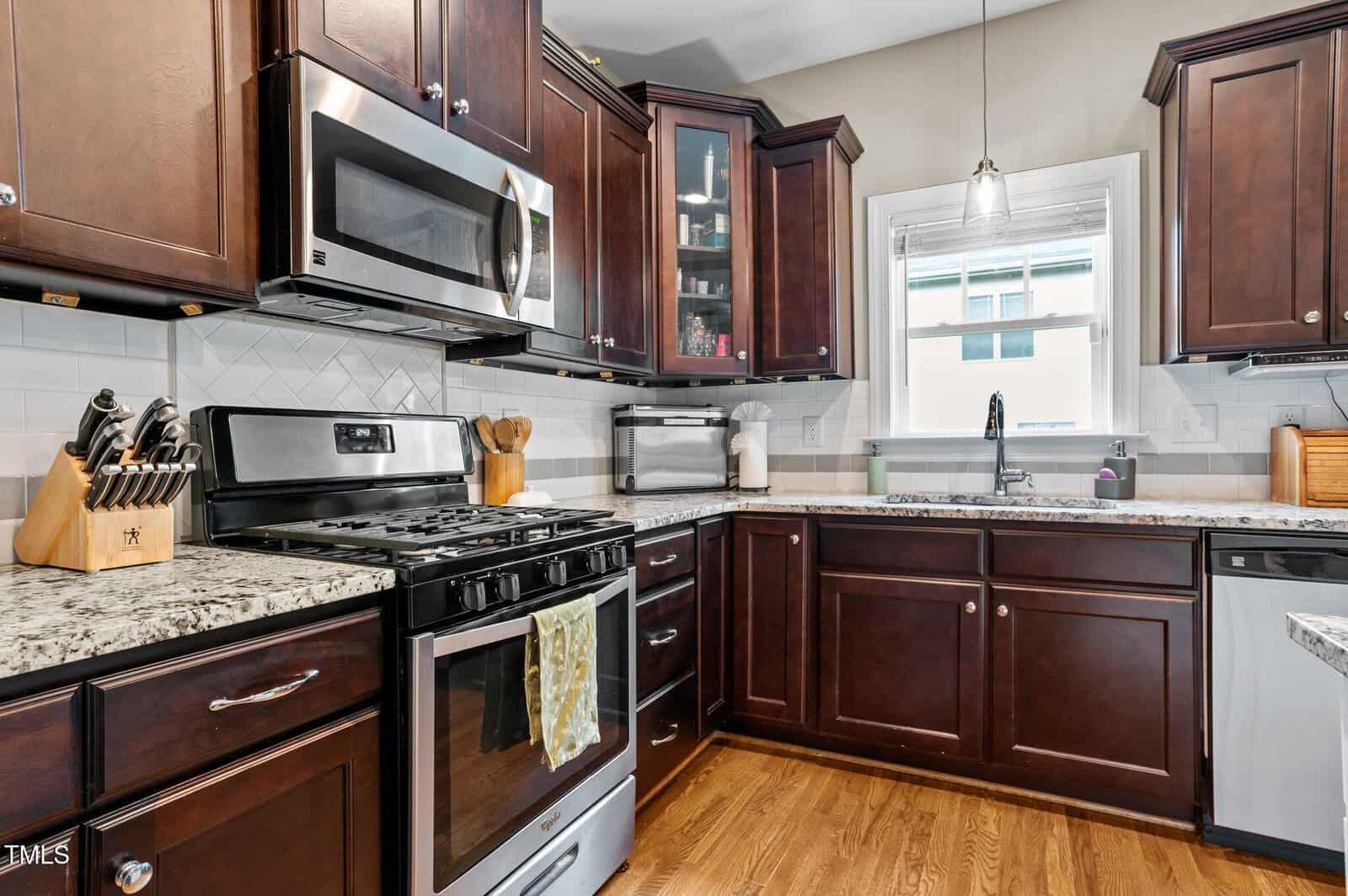 A modern kitchen with dark wood cabinets, stainless steel appliances, granite countertops, a knife block, and a window above the sink. There is a towel hanging on the oven and natural light coming through the window.