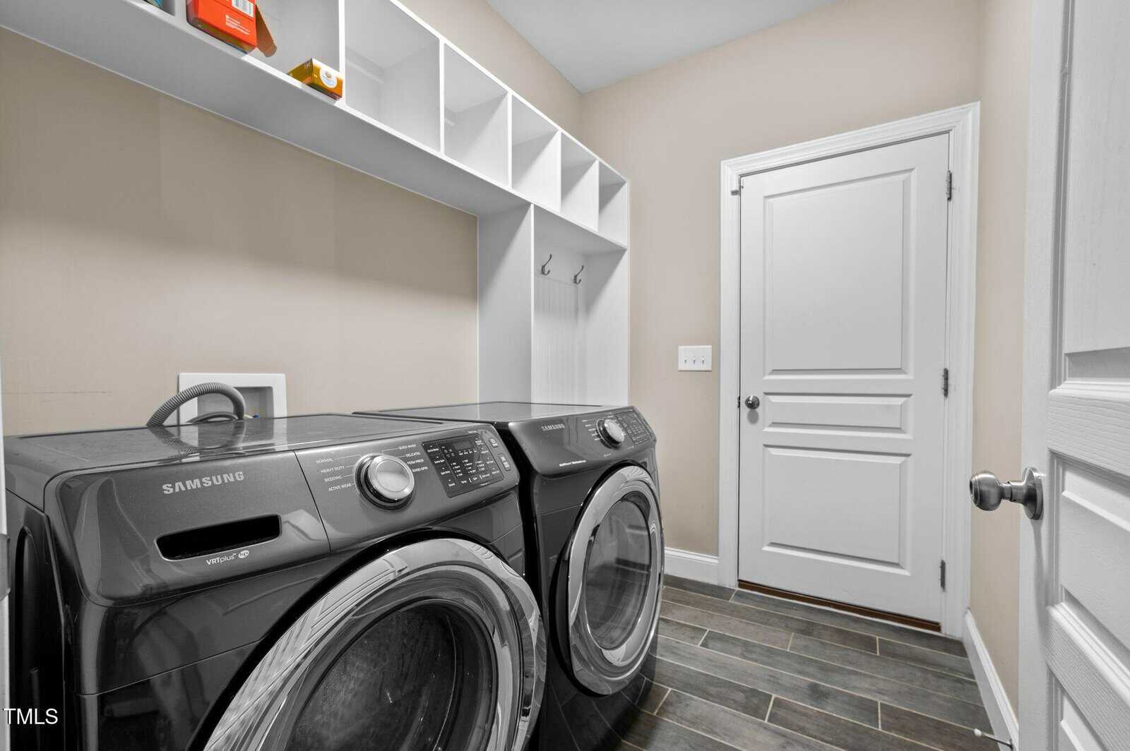 Laundry room with black Samsung washer and dryer, white shelves above, white walls, two white doors, and dark wood-look tile flooring. Hooks are mounted under the shelves for hanging items.