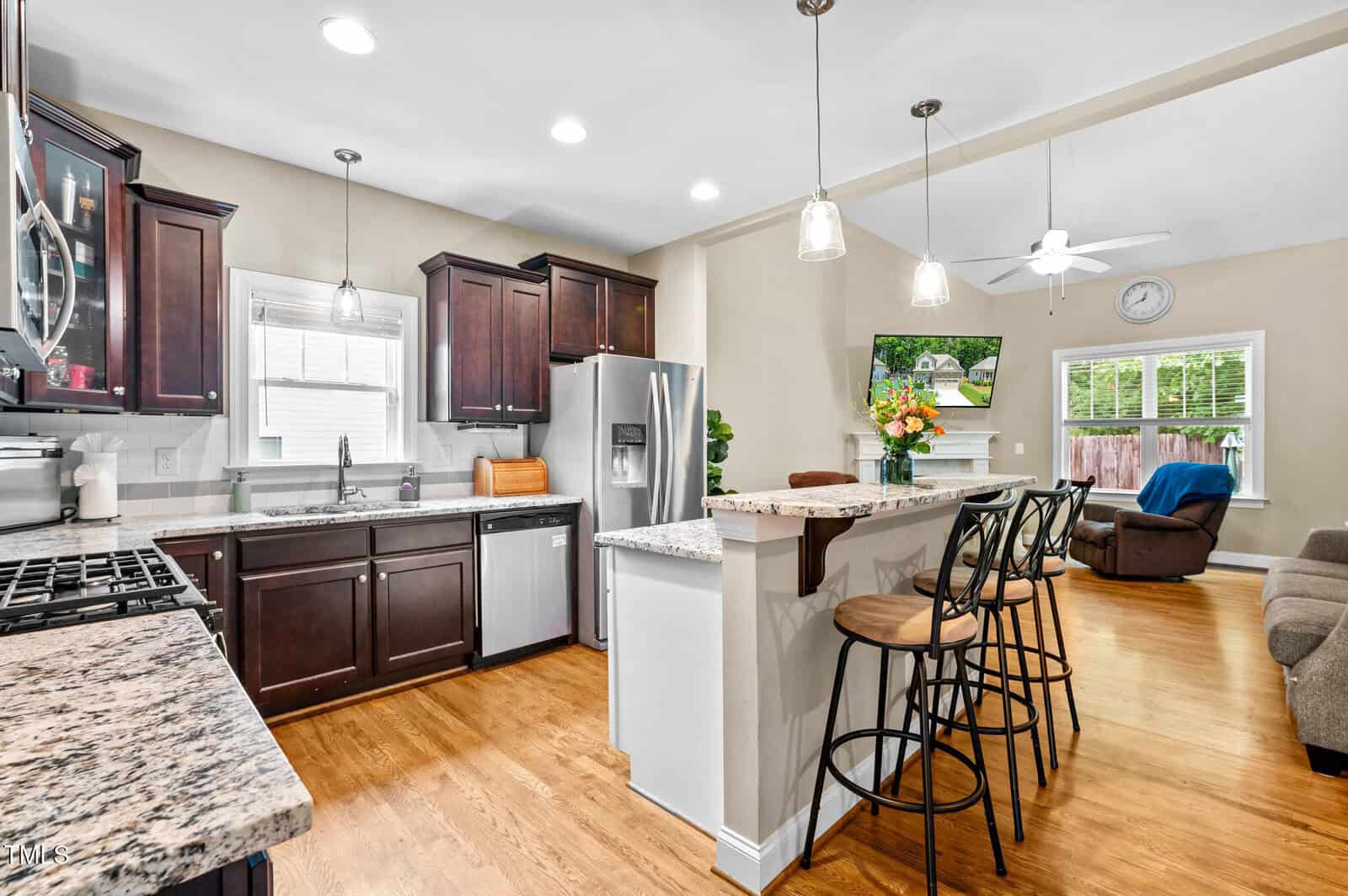 Modern kitchen with dark wood cabinets, stainless steel appliances, granite countertops, and a breakfast bar with three stools. Open layout leads to a living area with a sofa, recliner, wall art, and sliding glass doors.