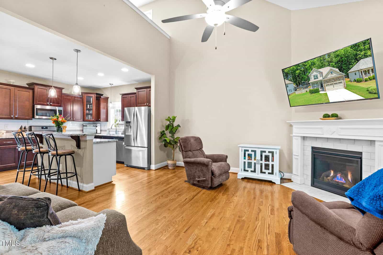 Open-concept living room and kitchen with hardwood floors, brown furniture, a white fireplace with a TV mounted above, stainless steel appliances, dark wood cabinets, and a breakfast bar with stools.
