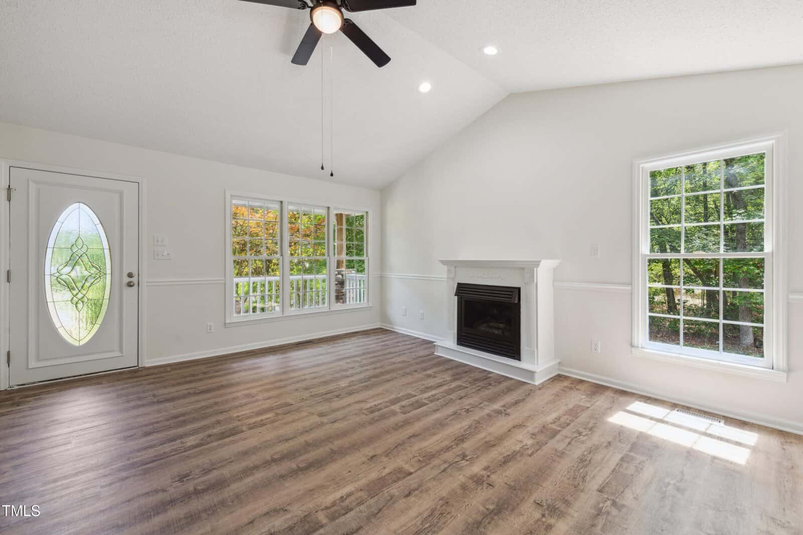 Bright living room with wood floors, a white fireplace, two large windows, a ceiling fan, and a glass door. Sunlight streams in, and trees are visible outside. The walls and ceiling are white.