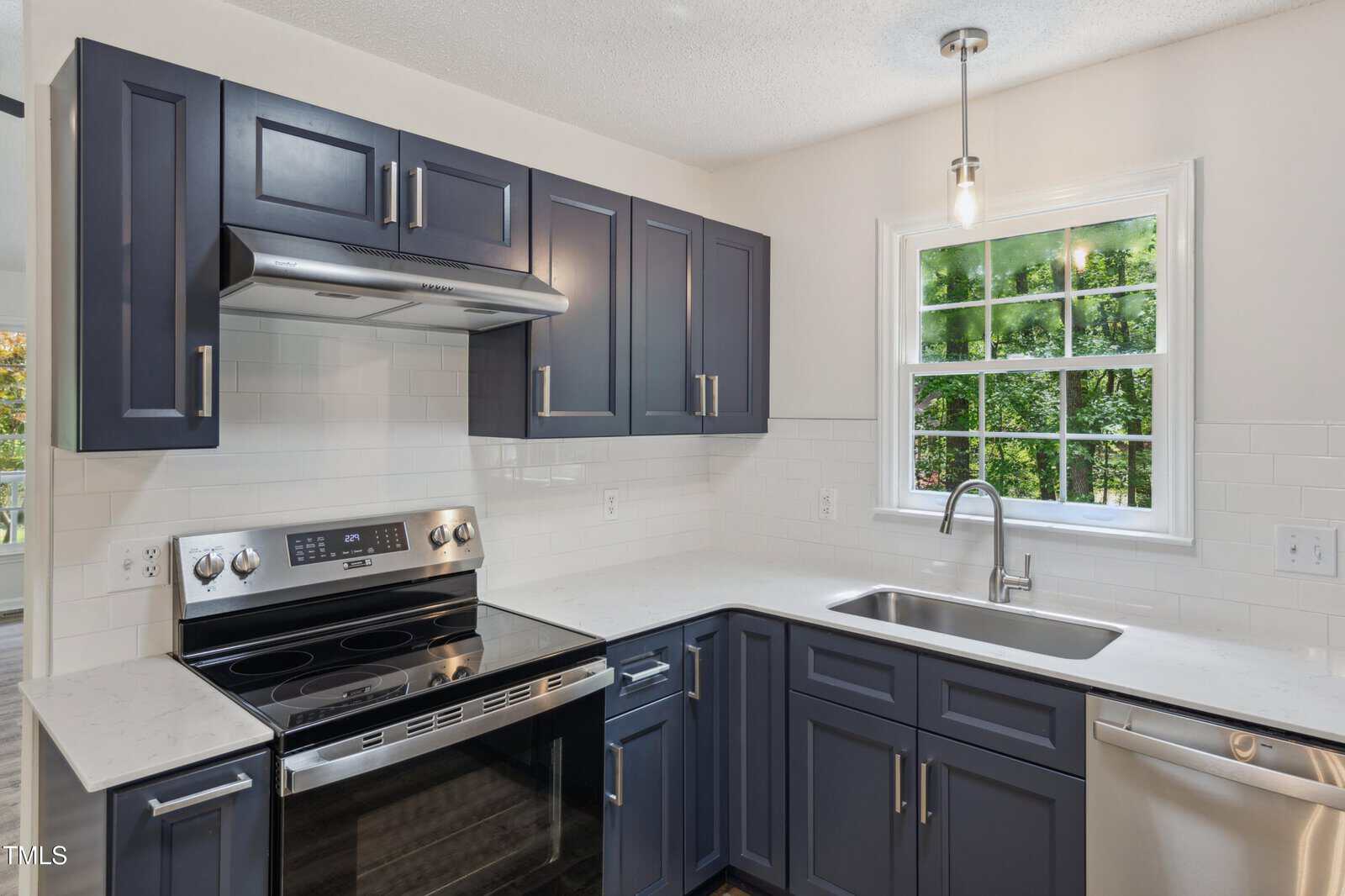 Modern kitchen with navy blue cabinets, stainless steel appliances, white subway tile backsplash, a single-basin sink under a window, and quartz countertops. Natural light streams through the window, illuminating the space.