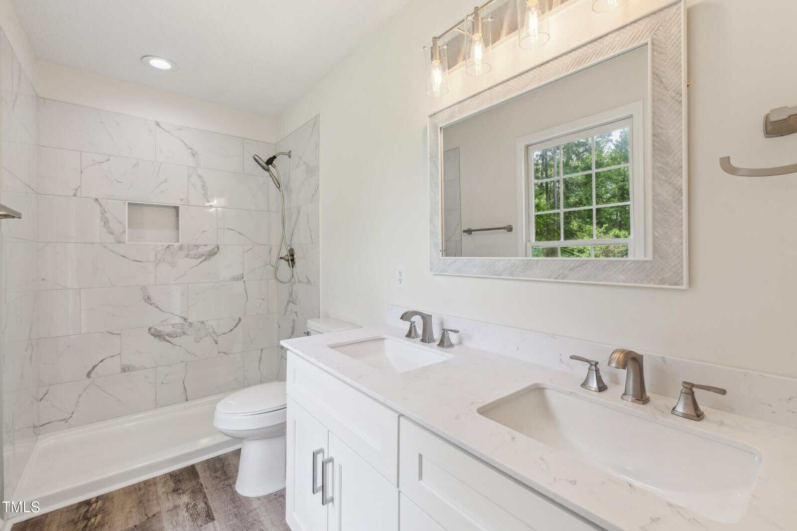 Modern bathroom with a white double-sink vanity, marble-style shower with built-in shelf, toilet, large framed mirror, three-light fixture, towel bars, and a window showing greenery outside.