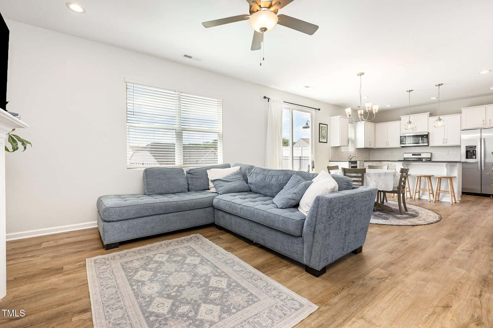 A modern living room with a blue sectional sofa, light patterned rug, ceiling fan, and wood floors, connected to a bright dining area and white kitchen with stainless steel appliances and pendant lights.