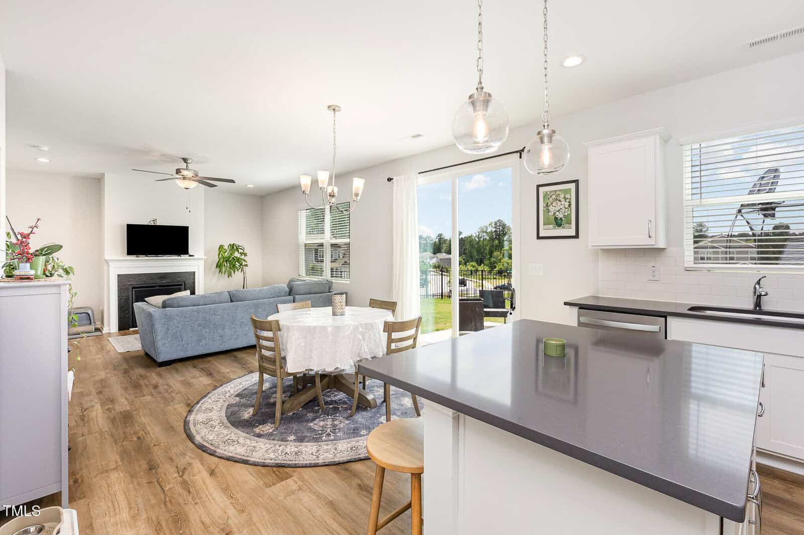 Modern open-concept living space with a kitchen, dining table, blue sectional sofa, fireplace, and large sliding glass doors leading to a backyard. Natural light fills the room through multiple windows.