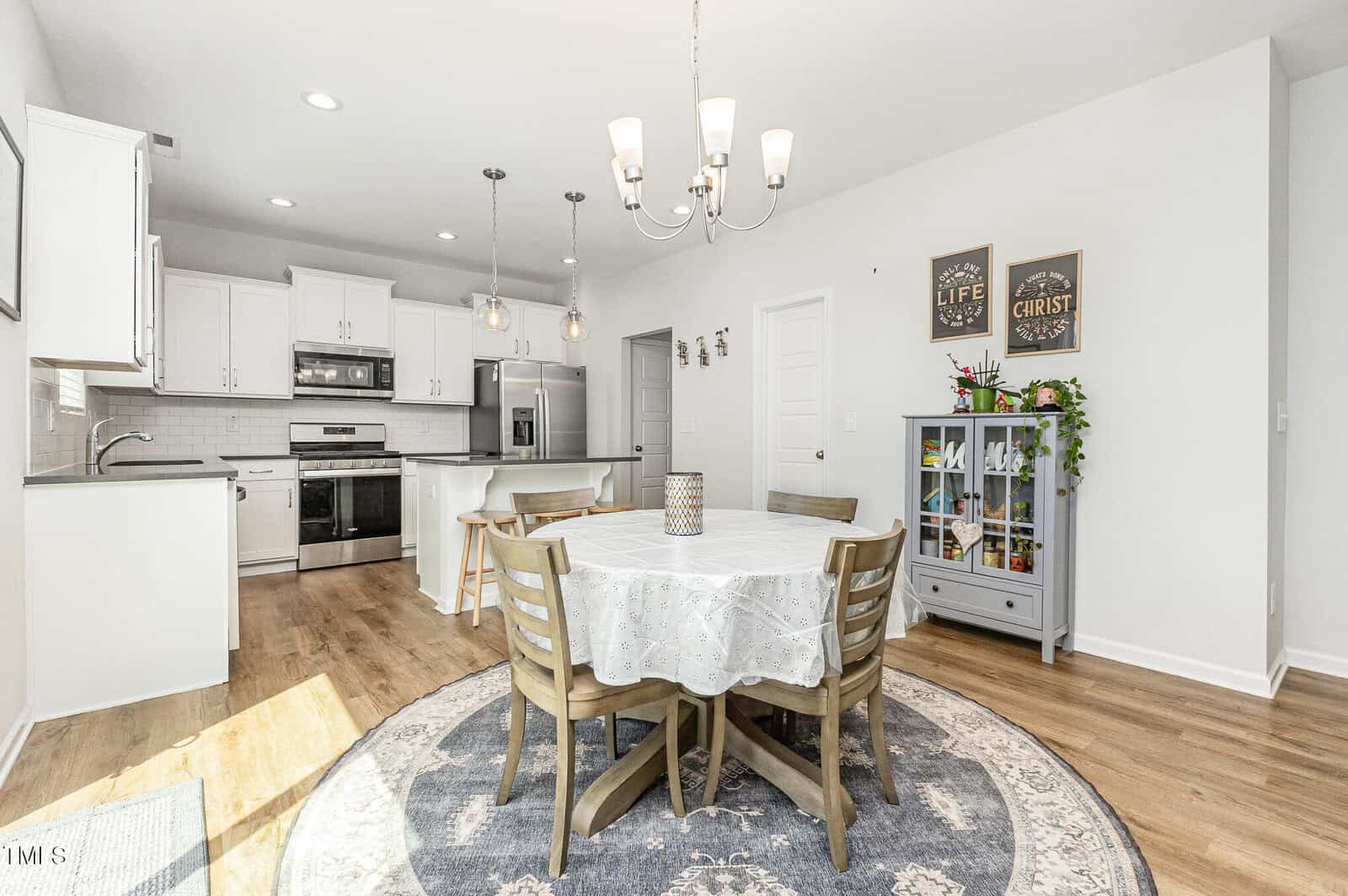 Bright kitchen and dining area with wooden floor, round table covered with a lace tablecloth, four chairs, gray side cabinet, and white cabinets. Pendant lights hang over the kitchen island. Wall decor and plants add accents.