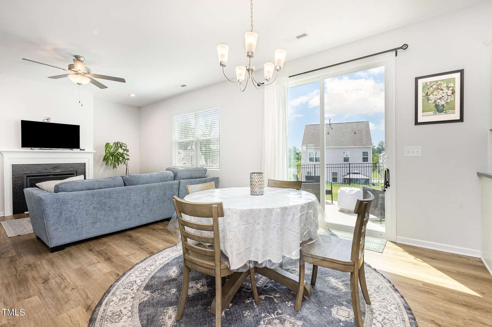 A bright dining area with a round table and four chairs on a rug, next to glass doors leading outside. A living room with a gray sofa, TV, and fireplace is visible. Hardwood floors and neutral walls complete the space.