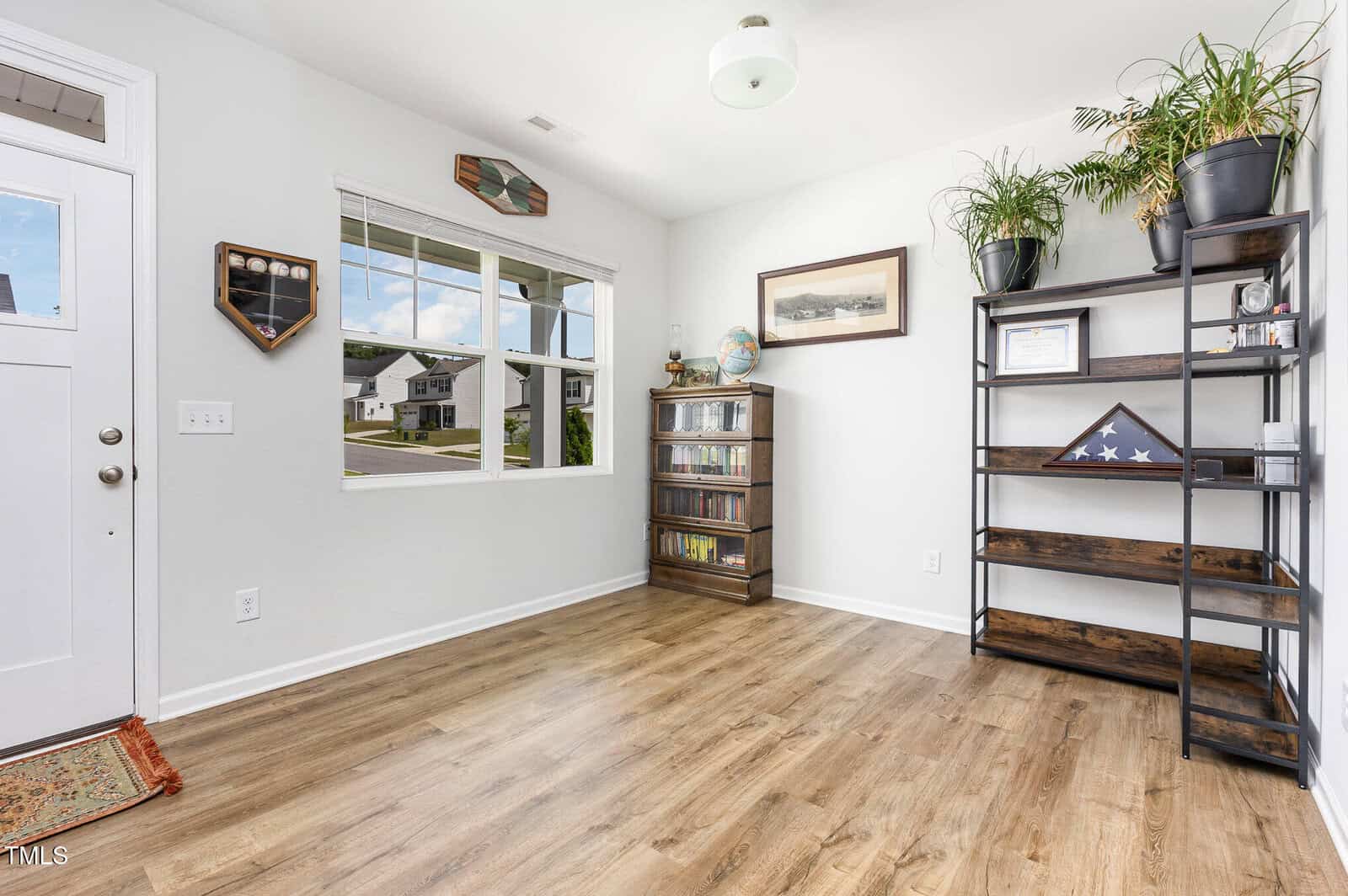 Bright home office with wood floors, large window, bookshelf with plants and memorabilia, and a CD cabinet under a framed picture. White walls and a partially visible door on the left.