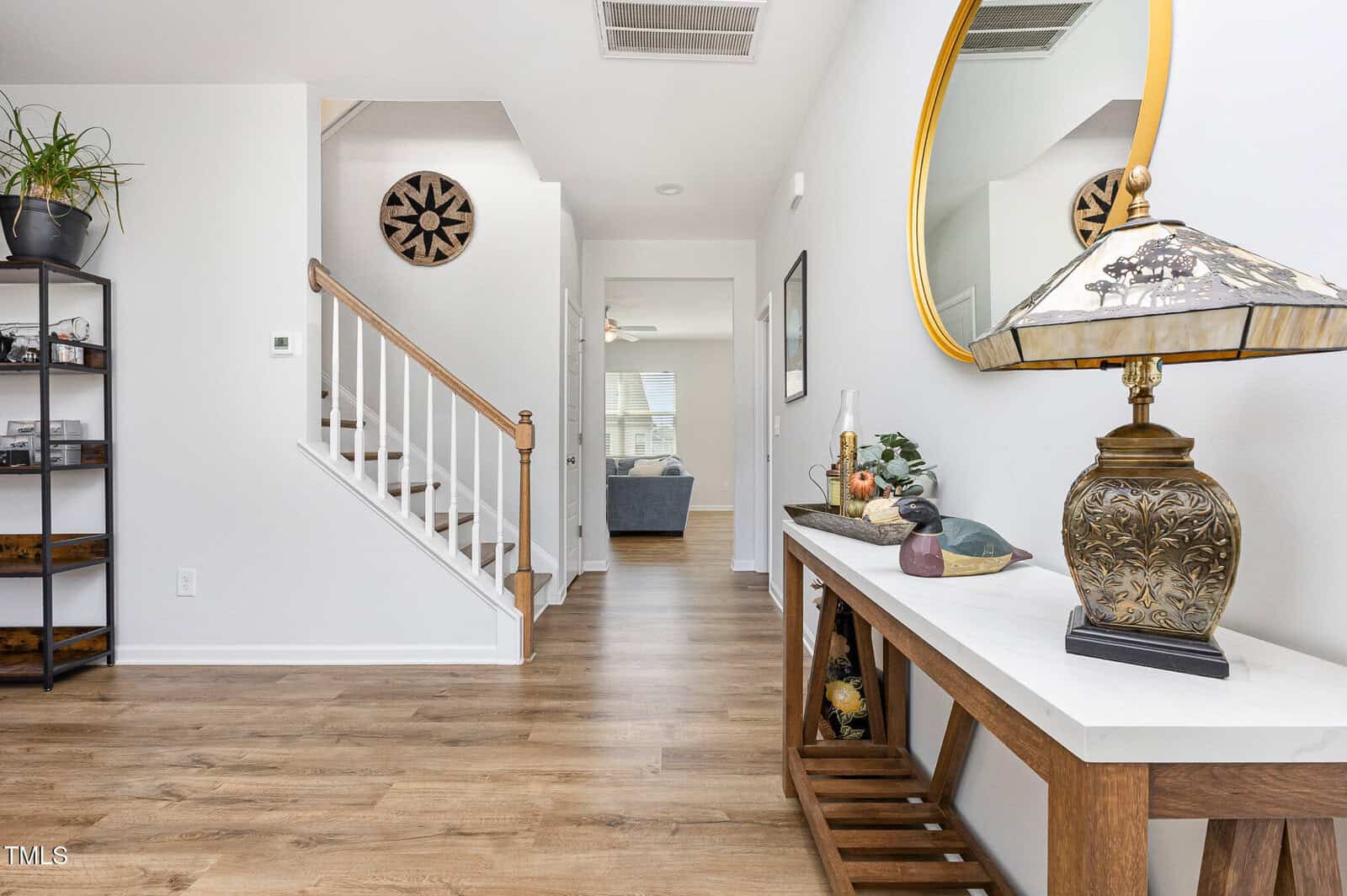 A hallway with wood flooring, a staircase with white risers and wood handrail, a console table with a decorative lamp and mirror, plants, and a glimpse into a living room with sofas in the background.
