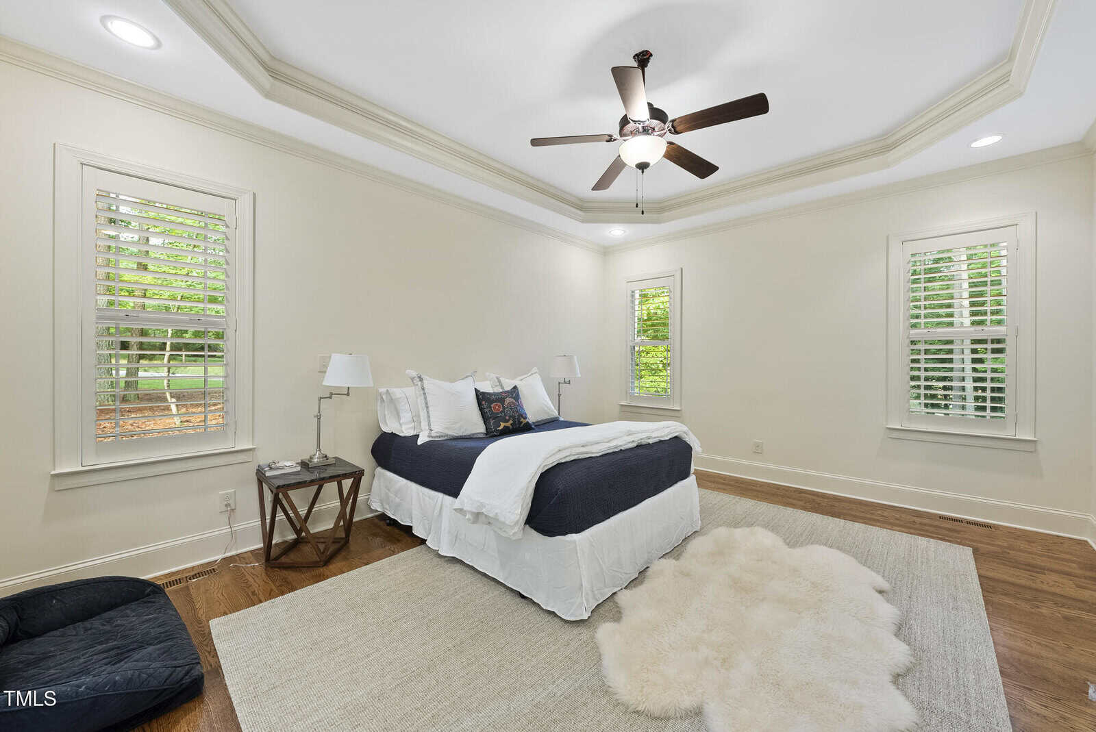 A bedroom with cream-colored walls, a ceiling fan, three windows with white shutters, a bed with white and dark bedding, two bedside lamps, a beige rug, a fur rug, and a pet bed on a hardwood floor.