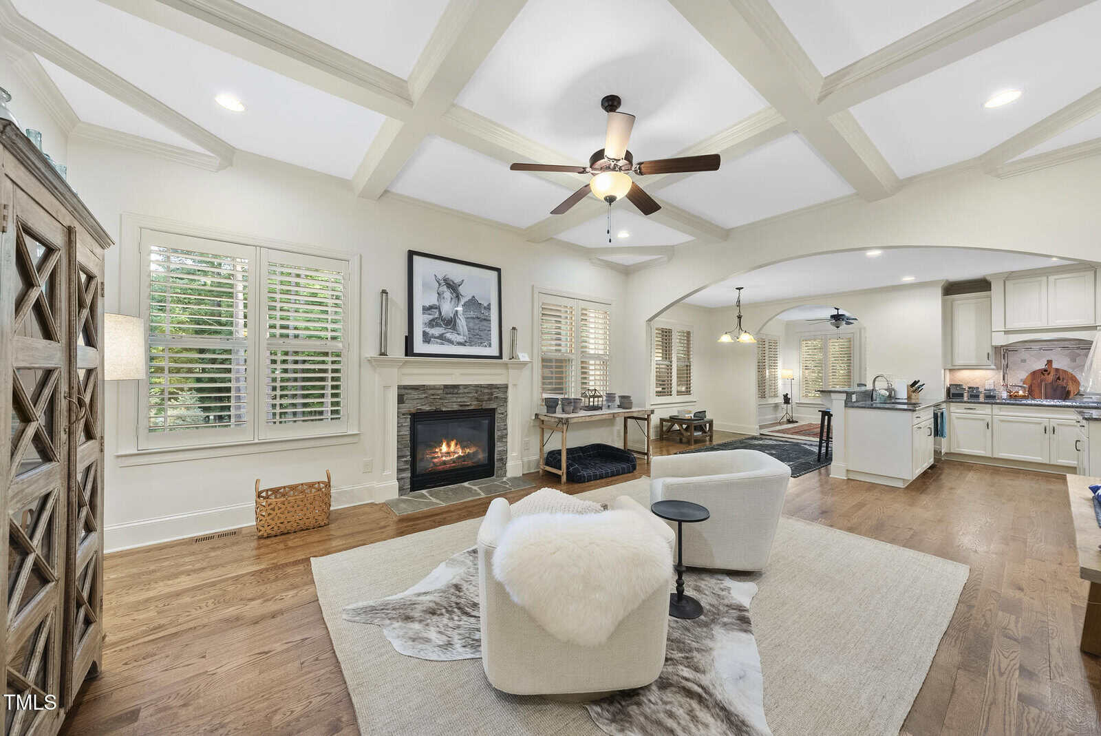 A bright living room with coffered ceiling, a fireplace, two light-colored chairs on a rug, wood floors, large windows with shutters, and an open view into the kitchen and dining area.