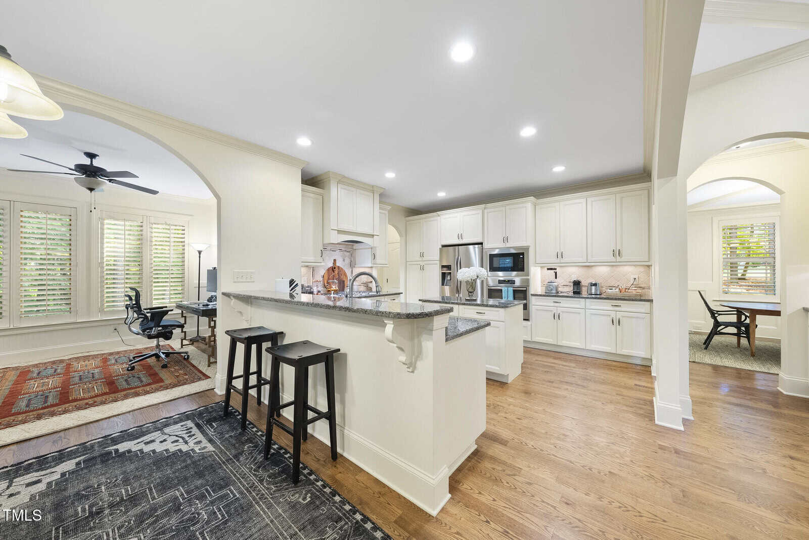 A spacious kitchen with white cabinets, stainless steel appliances, granite countertops, and a breakfast bar with two black stools. Hardwood floors and multiple archways open to adjacent rooms with natural light.