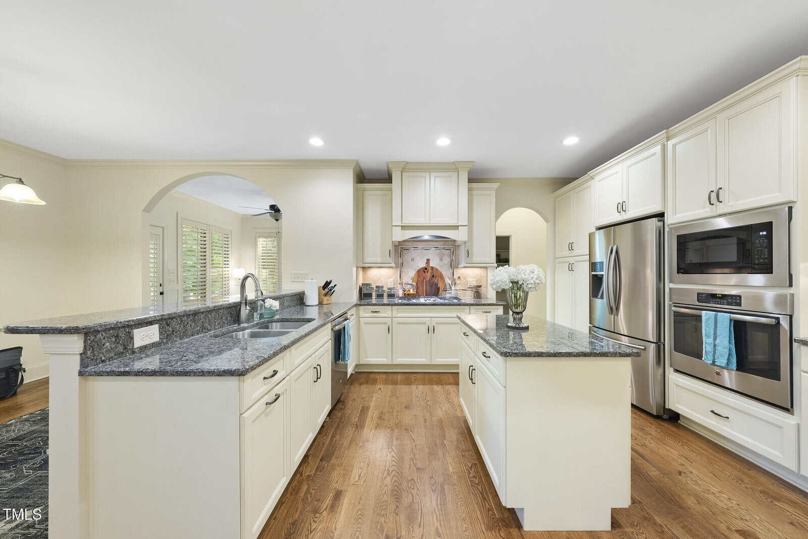 Spacious kitchen with white cabinets, stainless steel appliances, granite countertops, a center island with a vase of flowers, wood flooring, and recessed lighting. An archway leads to another room.
