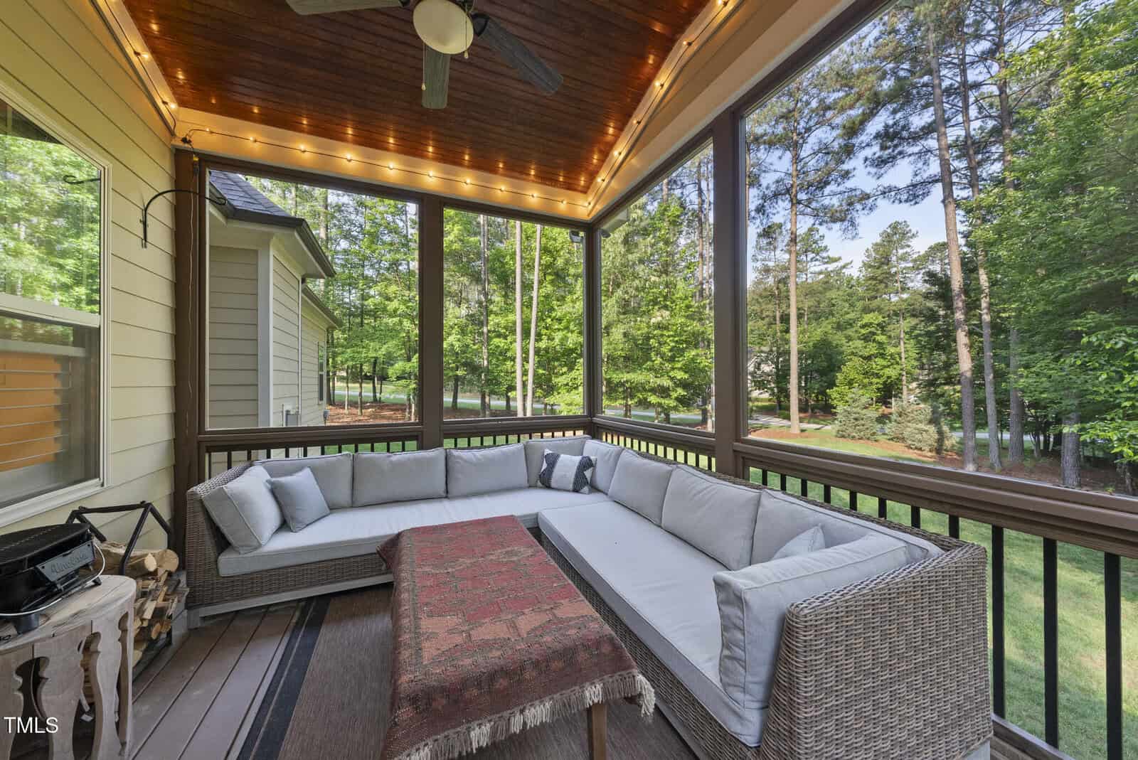 A screened-in porch with a wooden ceiling, string lights, a ceiling fan, a large L-shaped gray outdoor sofa with cushions, a red patterned rug on the coffee table, and views of a green backyard with trees.