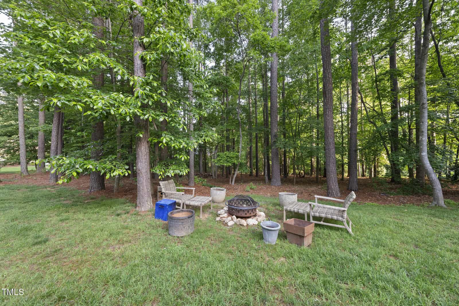 A wooded backyard area with tall trees, a fire pit surrounded by benches and chairs, and various containers and a cardboard box on the grass. The setting is shaded and the ground is covered with green grass.