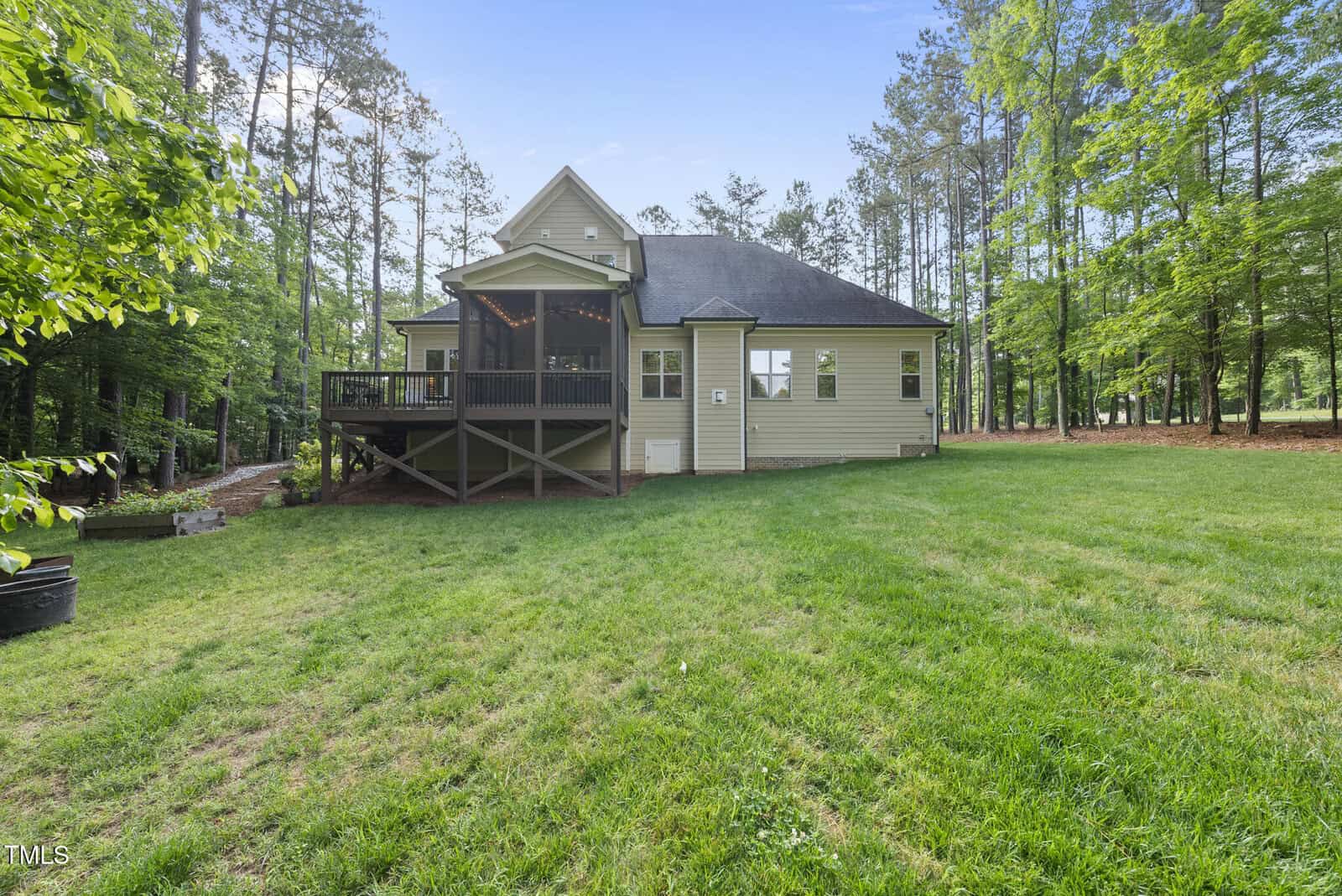 Single-story house with light-colored siding, black roof, and a screened-in back porch, situated on a large grassy yard surrounded by tall trees. The area appears open and well-maintained.