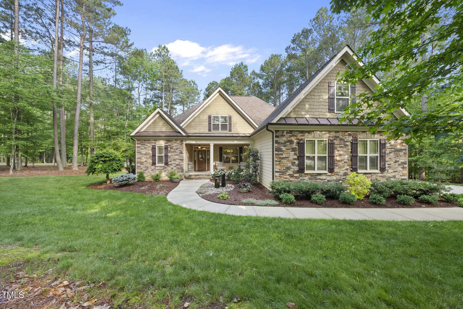 Single-story house with stone and wood siding, gabled roof, front porch, manicured lawn, shrubs, and tall trees in the background under a clear blue sky.