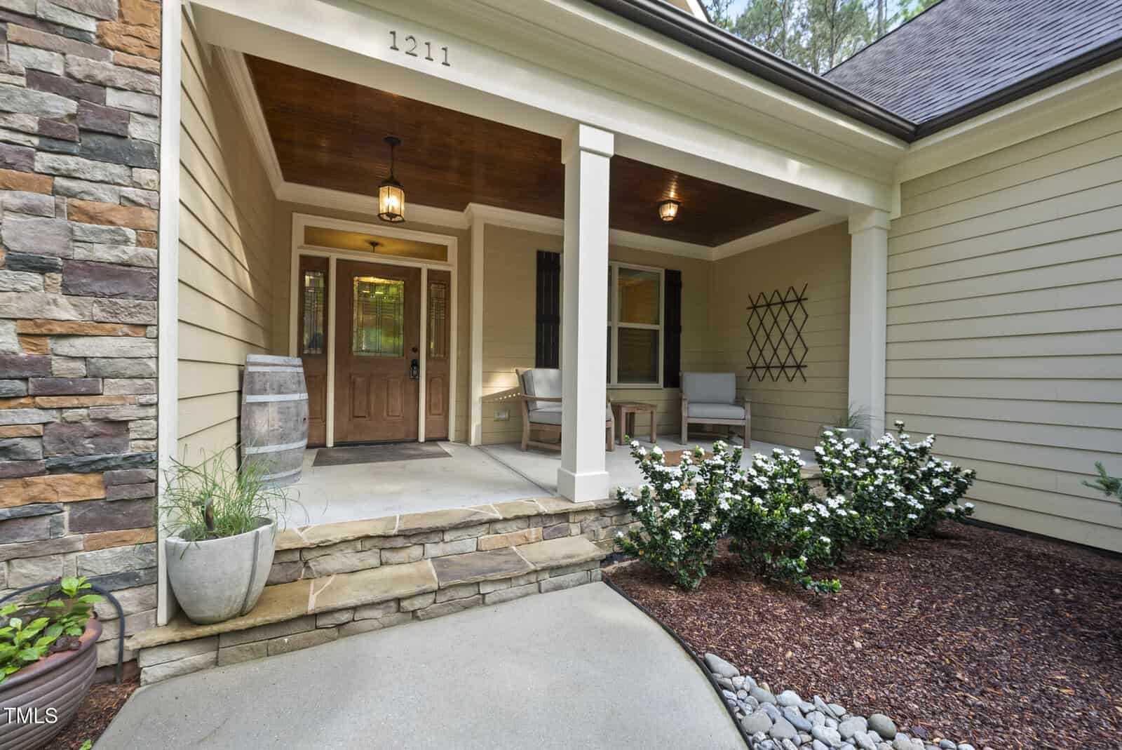 A cozy front porch with a wooden door, two white chairs, potted plants, a barrel, and white blooming bushes. The house exterior features beige siding, stone accents, and a wooden ceiling. House number 1211 is above the door.