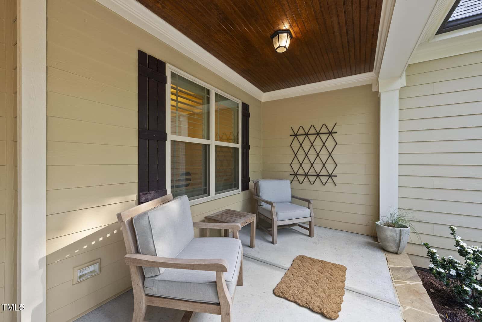 Covered front porch with two cushioned wooden chairs, a small wooden table, a brown doormat, potted plants, and wall decor. The ceiling is wood-paneled with a light fixture.