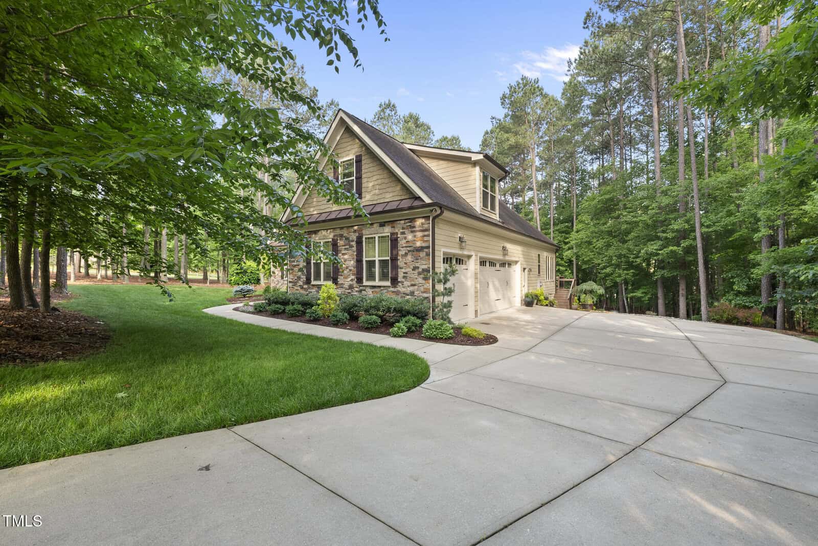 A two-story house with stone and siding exterior, white garage doors, and landscaped front yard, surrounded by tall trees and a wide concrete driveway curving to the entrance.