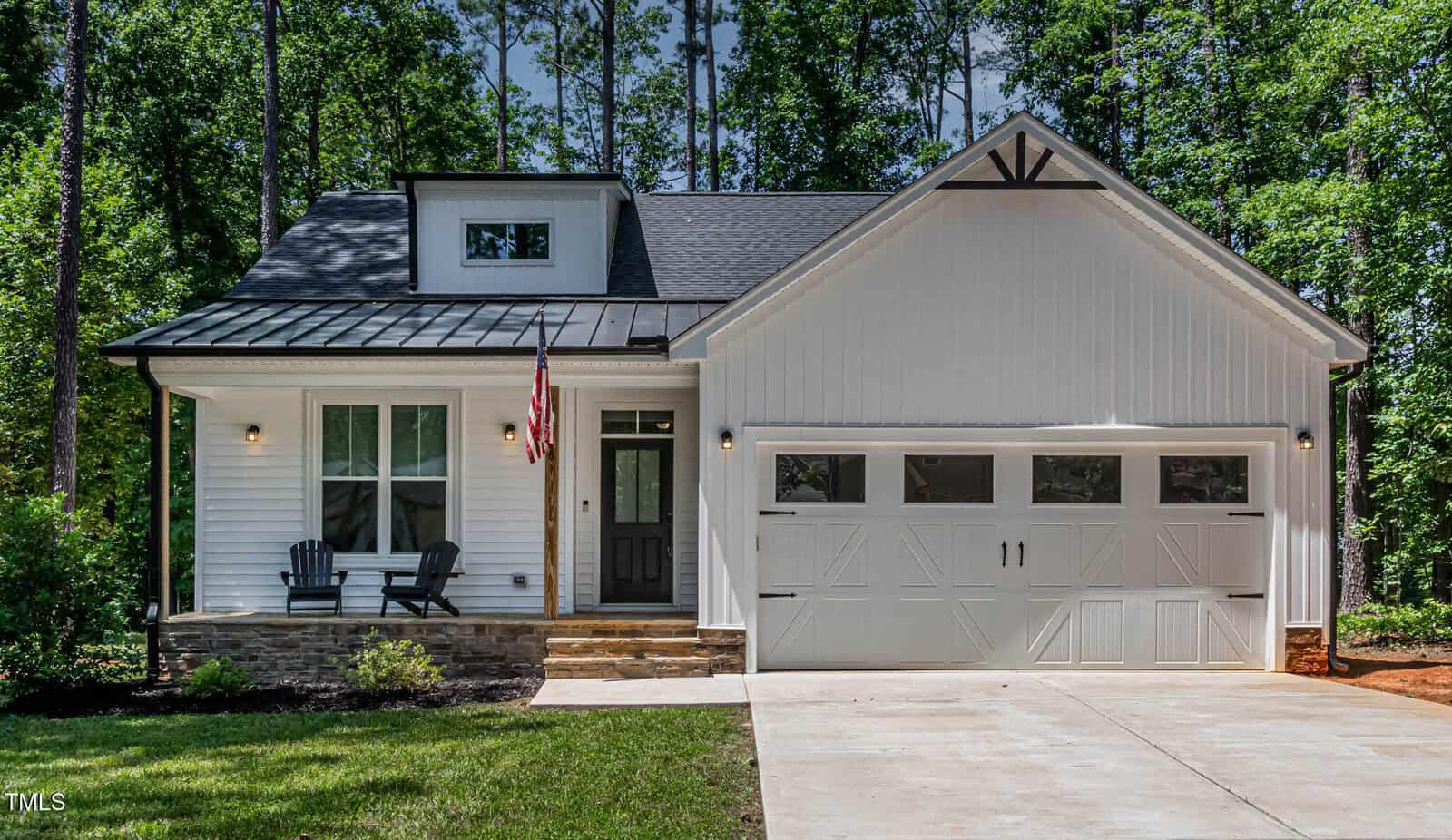 A modern white house with a black roof, a two-car garage, front porch with two chairs, and an American flag. The house is surrounded by green grass, shrubs, and tall trees.