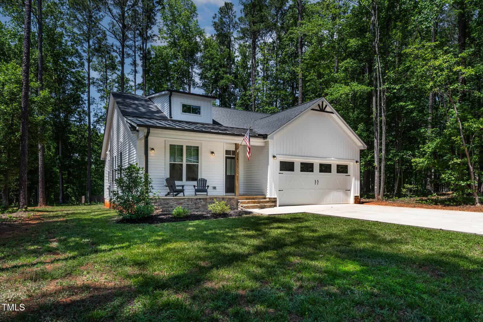 A modern white single-story house with a double garage, front porch with chairs, American flag, and surrounded by tall trees and greenery on a sunny day.