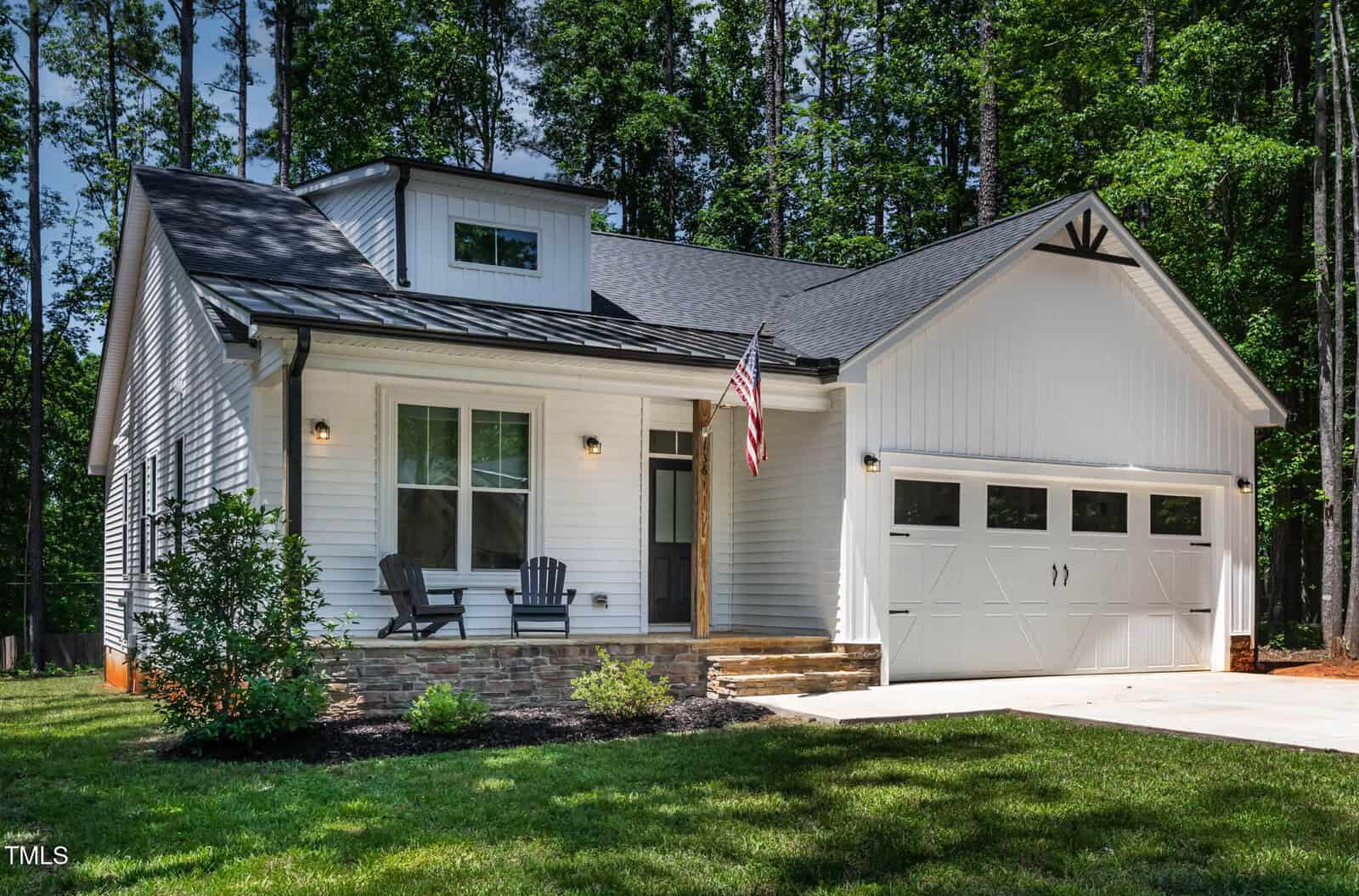 A modern white house with a gabled roof, attached two-car garage, front porch with two black chairs, American flag, and a manicured lawn, set against a backdrop of tall trees.