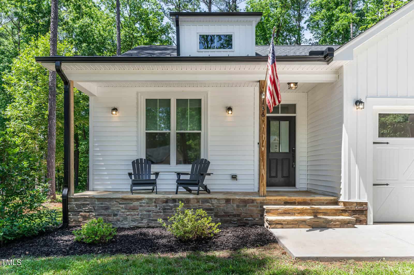 A white house with a stone porch, two black chairs, an American flag, and black-trimmed windows, surrounded by greenery and trees. There is a sidewalk leading to the front door.
