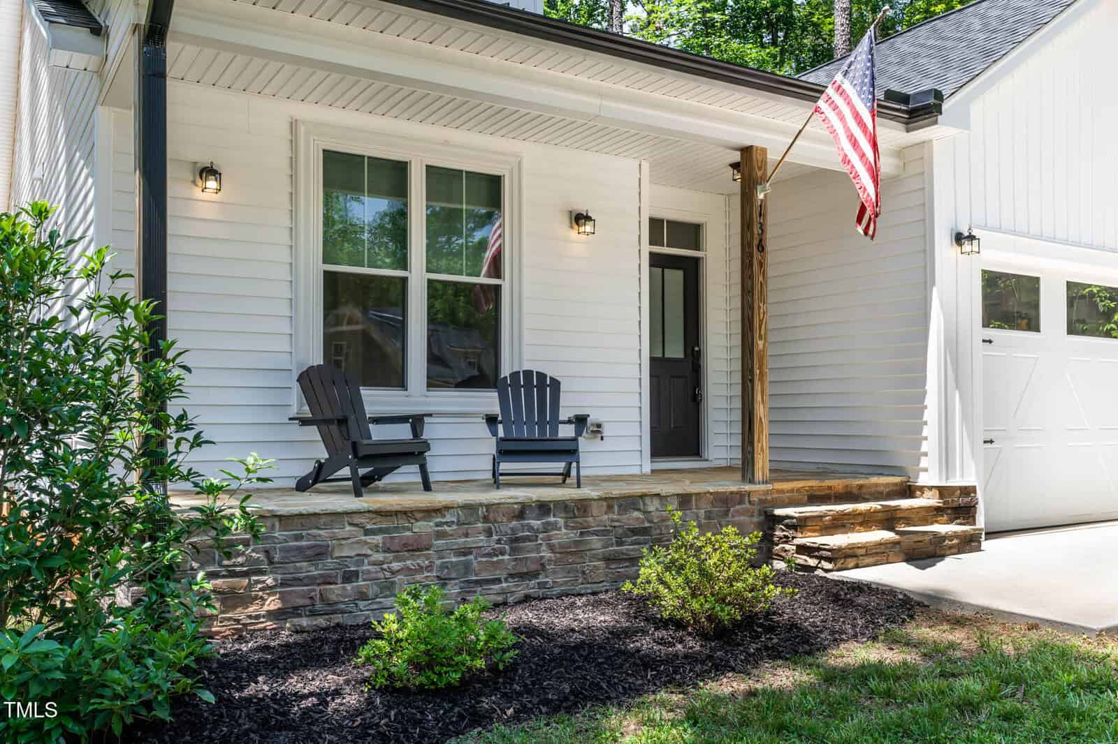 White house with stone porch, two black chairs, American flag, front door with sidelight, exterior lights, white garage door, and landscaped mulch area with green bushes in front.