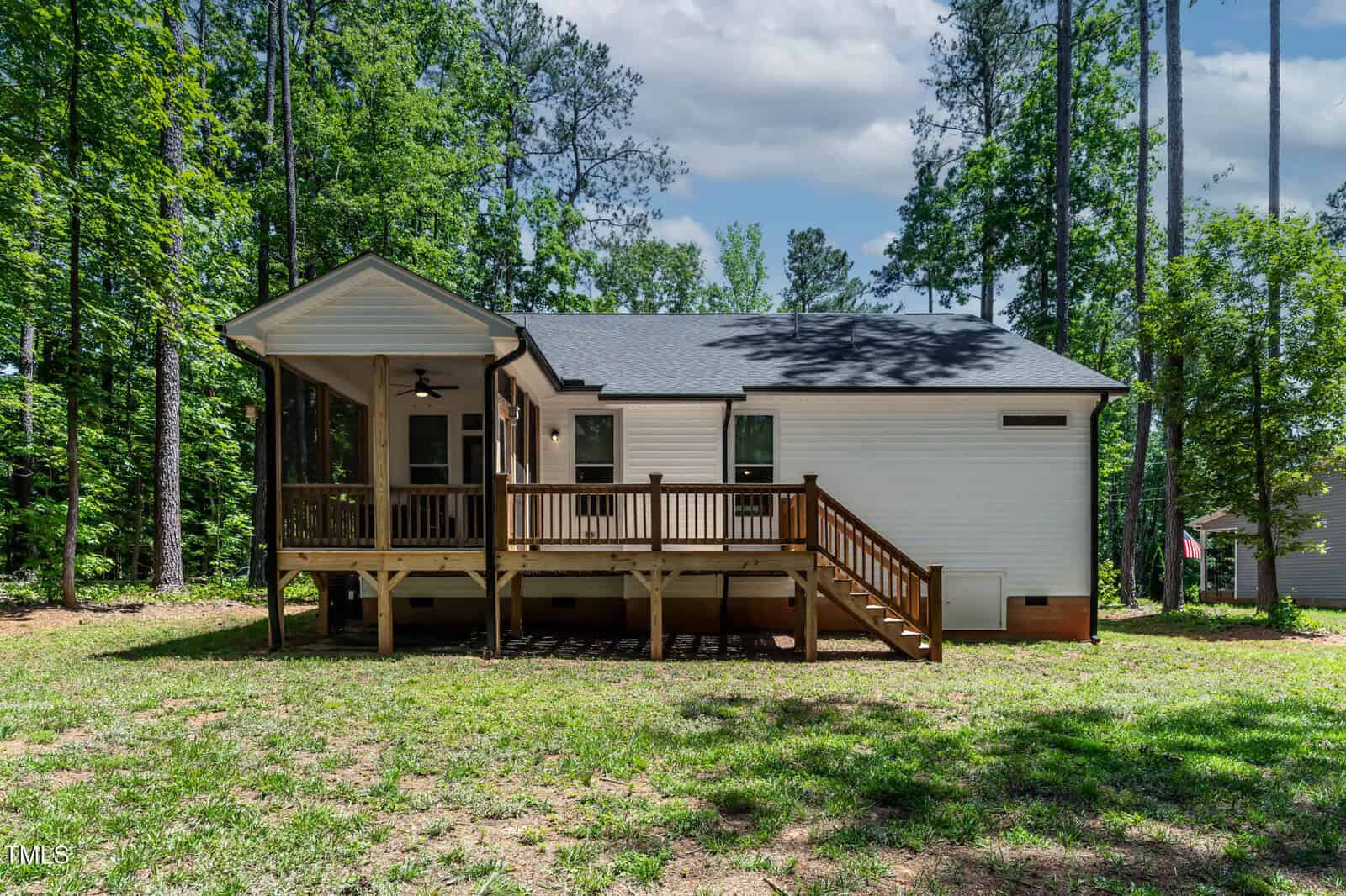 A single-story white house with a black roof, wooden deck, screened porch, and steps, surrounded by trees and grass under a partly cloudy sky.