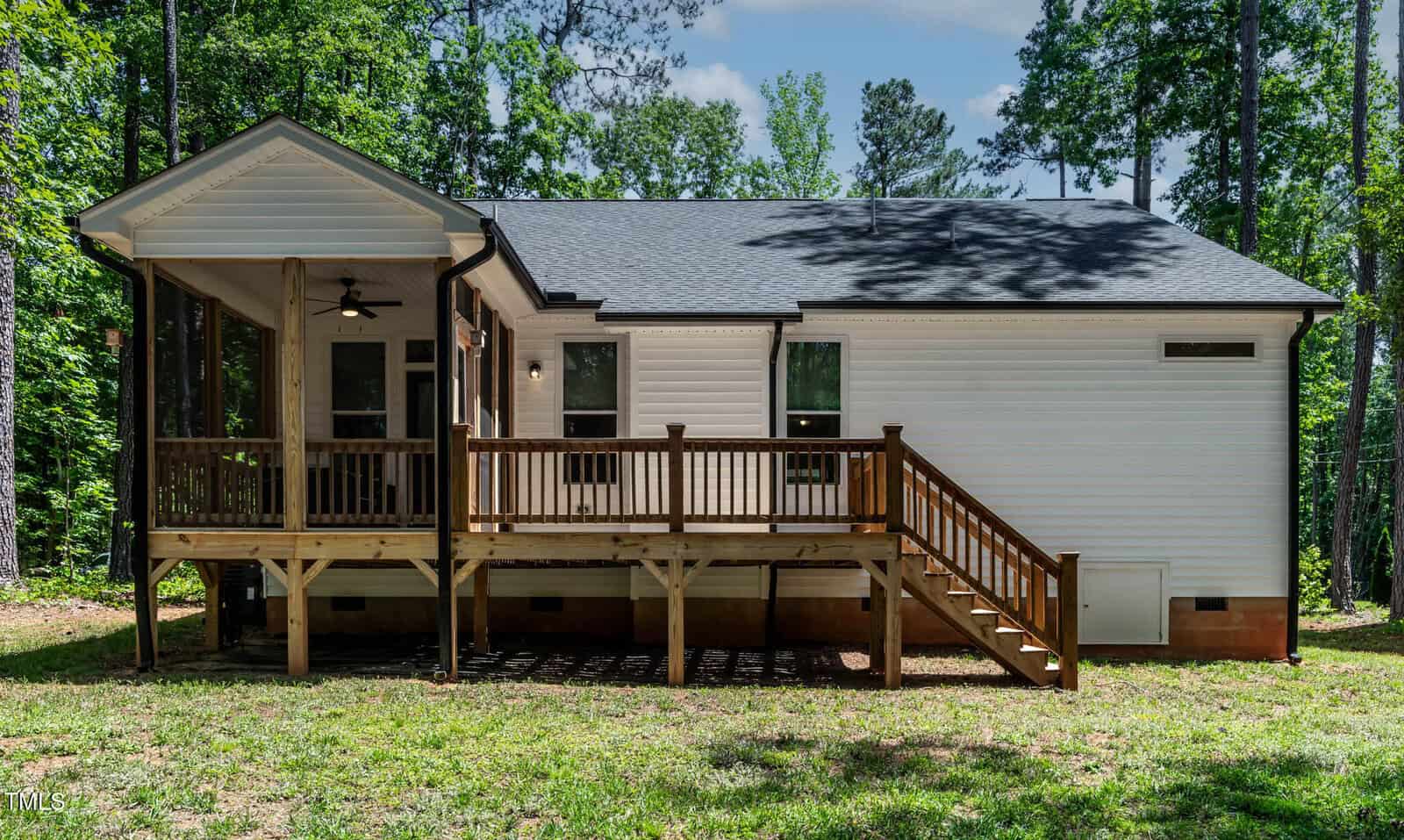 Single-story white house with a black roof, a wooden deck, and stairs leading to a grassy yard. The house is surrounded by tall trees and stands on a raised foundation. A screened-in porch is visible on the left side.