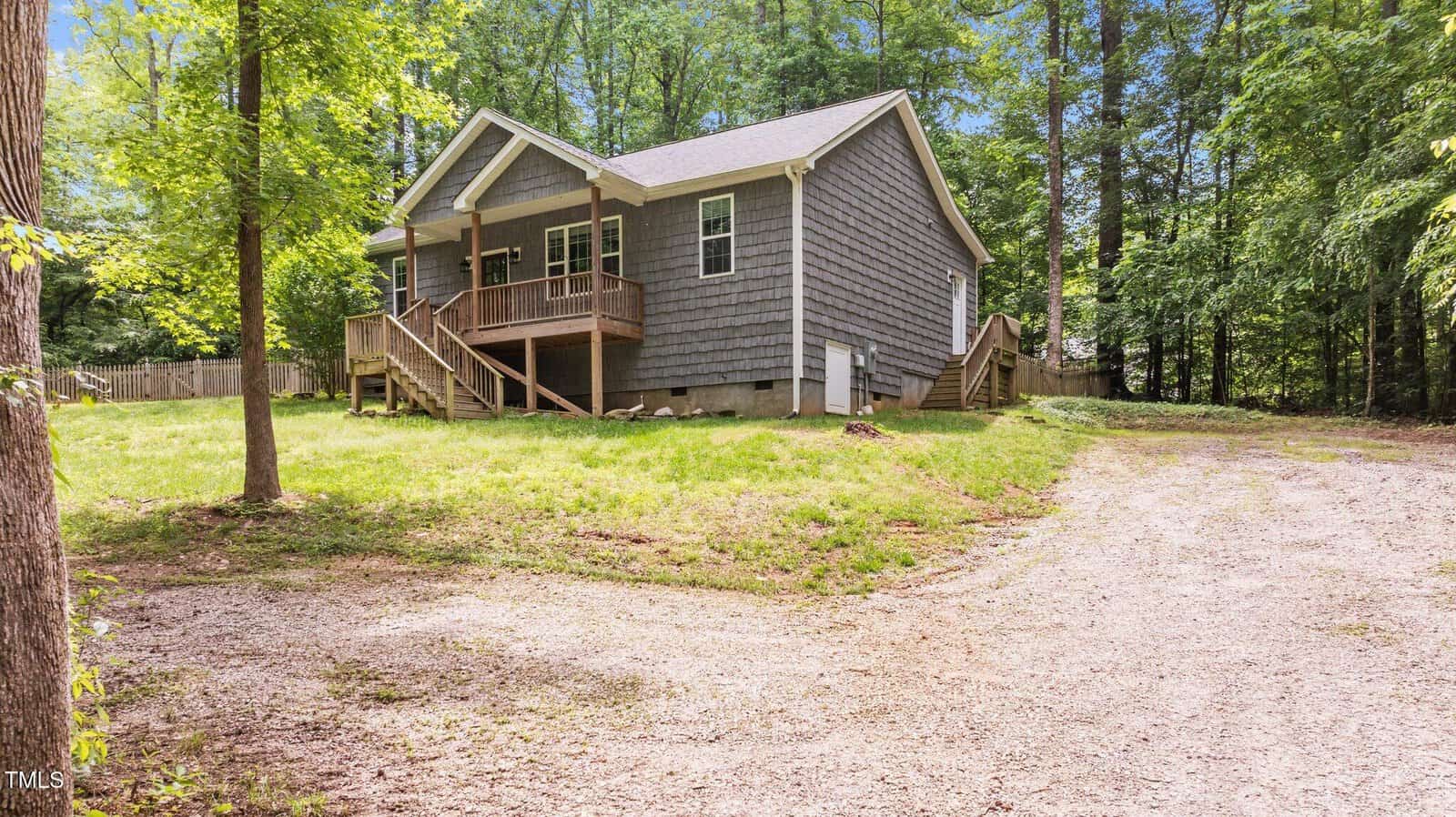 A small gray house with a front porch and steps, surrounded by green grass and tall trees, sits next to a curved gravel driveway in a wooded area on a sunny day.