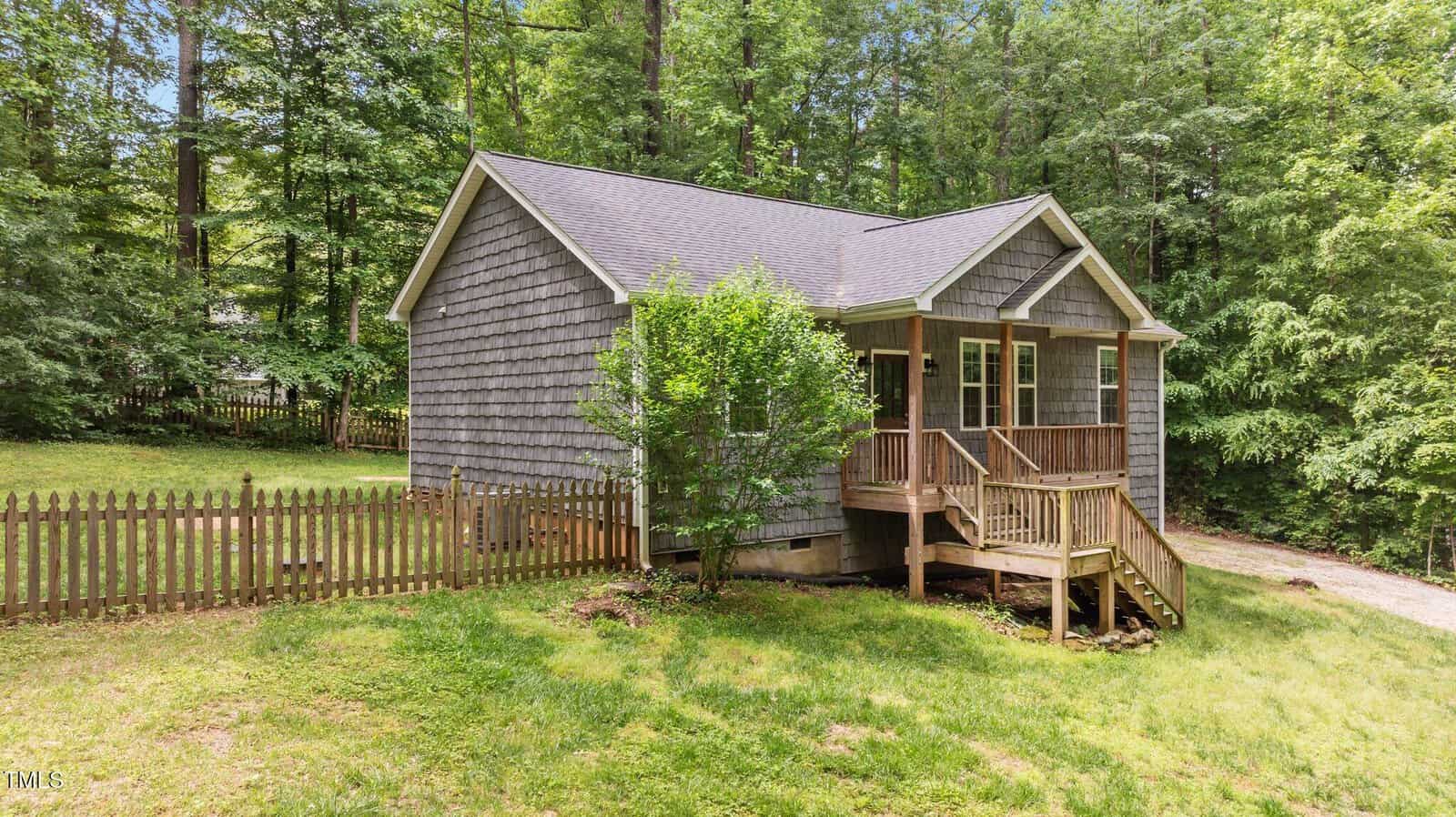 A small wooden cabin with a front porch and steps, surrounded by a picket fence and lush green trees in a forested area.