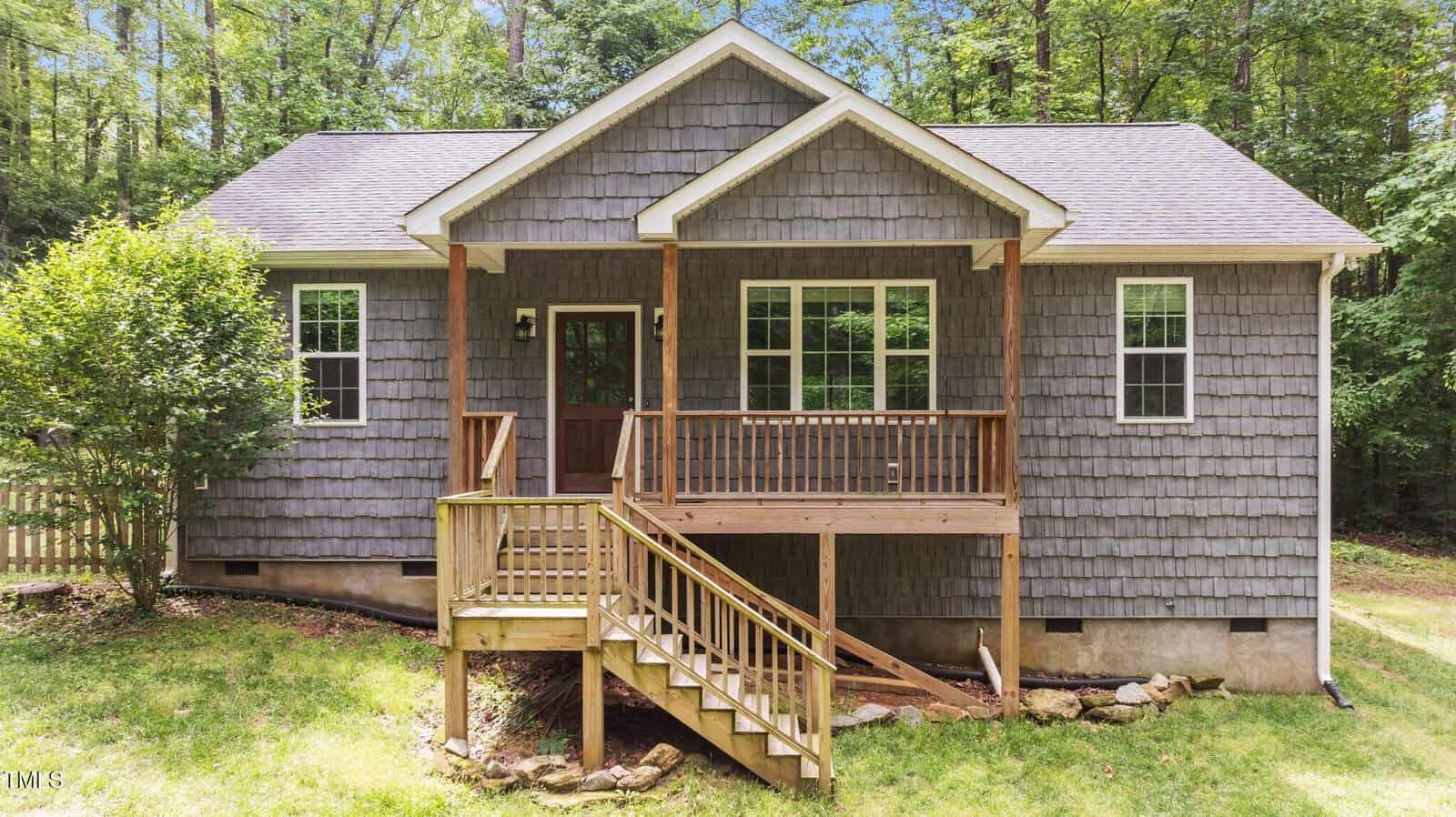 A small gray house with wooden shingles, a gabled roof, and a front porch with stairs, set in a grassy yard and surrounded by trees.