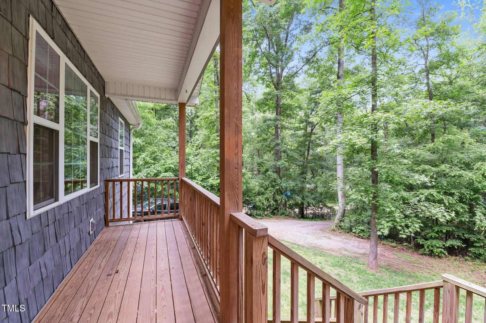 A wooden porch with a railing looks out onto a wooded area with green trees and a gravel driveway under a blue sky.