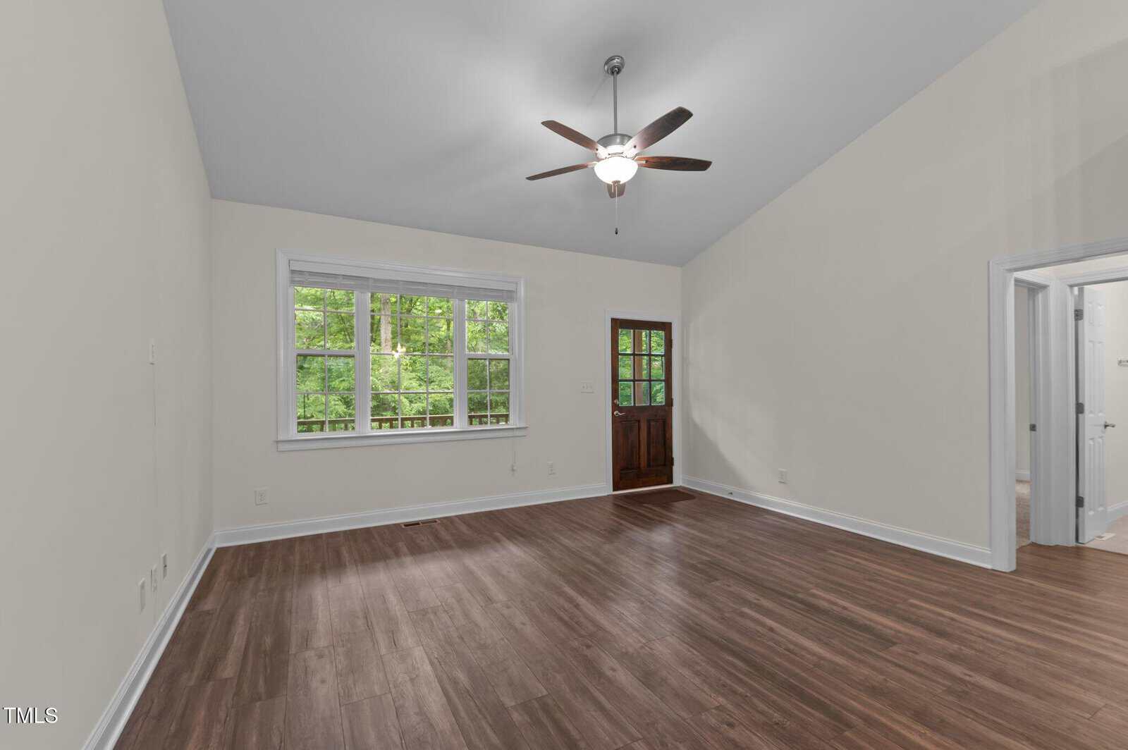 Empty room with beige walls, wood flooring, a ceiling fan with light, three windows, and a wooden door. The room receives natural light from the windows, and a doorway leads to another area.