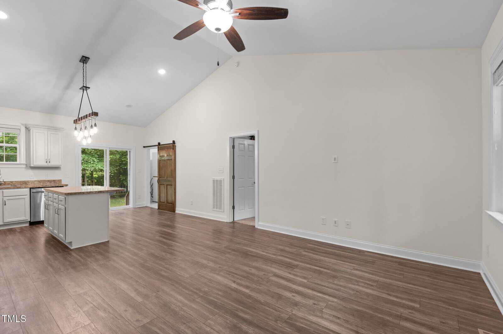Open living space with wood flooring, white walls, vaulted ceiling with fan, sliding barn door, modern light fixture, and a kitchen with white cabinets, island, and windows overlooking greenery.