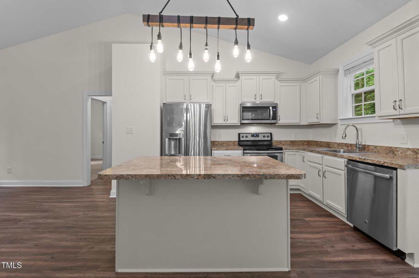 Modern kitchen with white cabinets, stainless steel appliances, brown granite countertops, island with overhead rustic light fixture, and dark wood floors. Window above sink lets in natural light. Doorway leads to another room.
